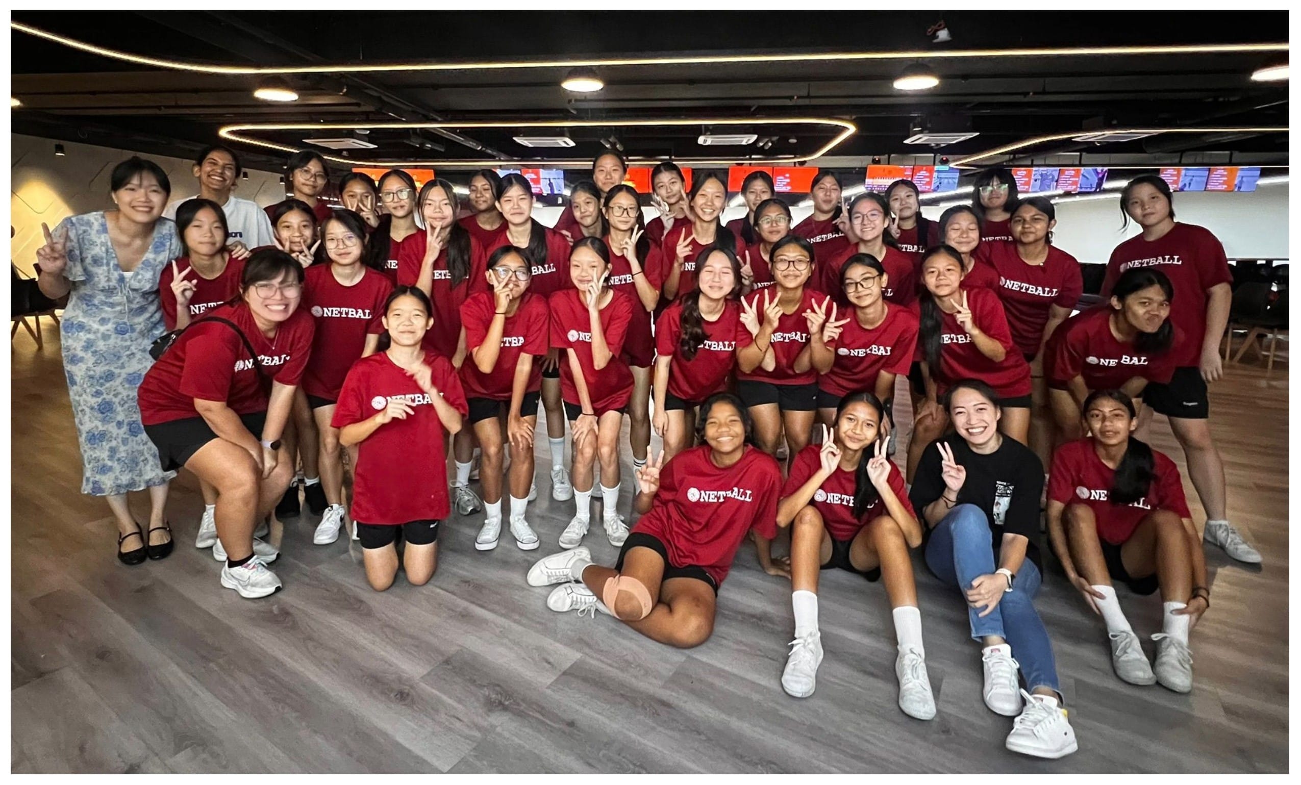 Large group of young women in red Netball shirts posing with peace signs indoors on a wooden floor.
