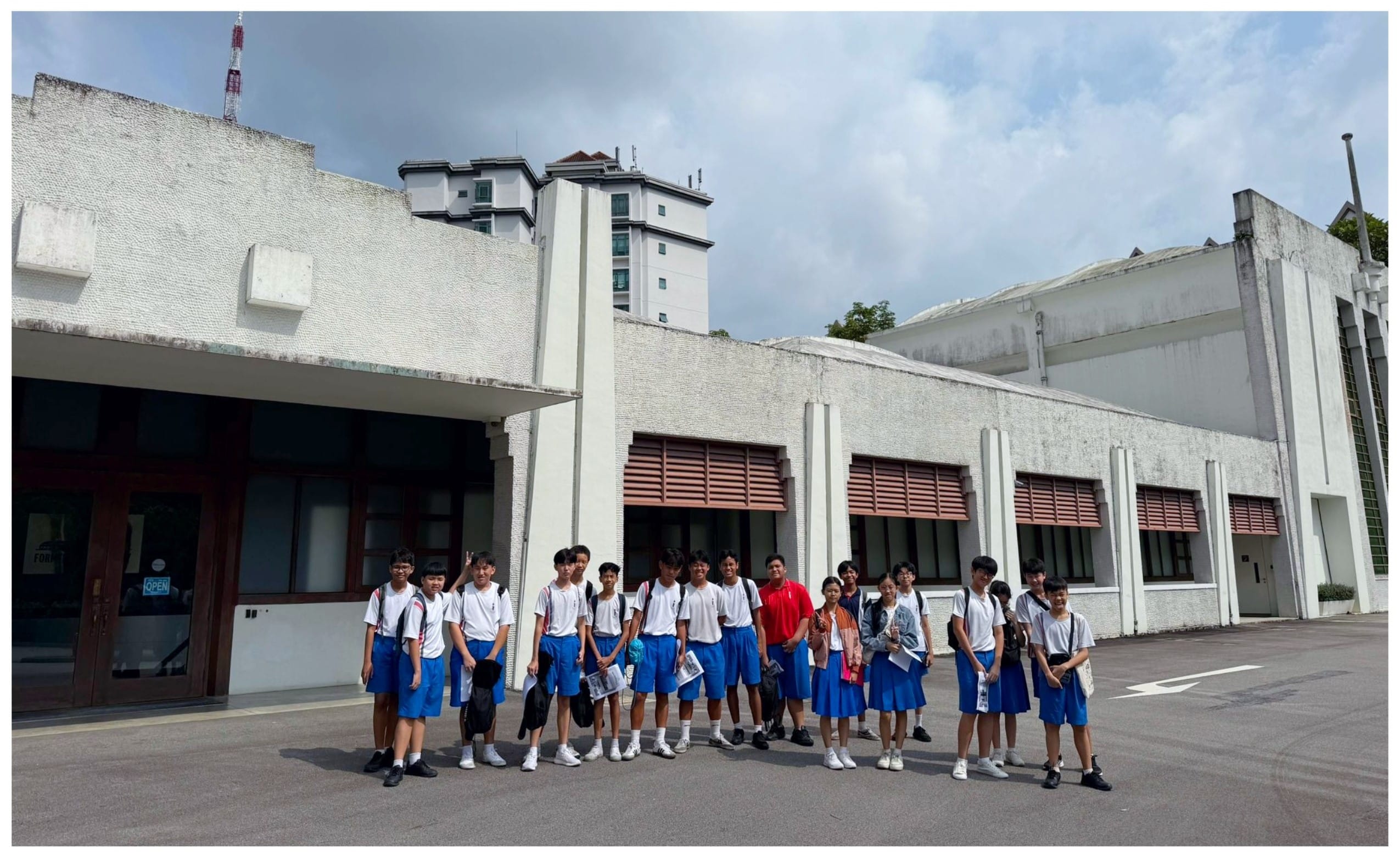 A group of students in white shirts and blue uniforms stand together outside a large white building.