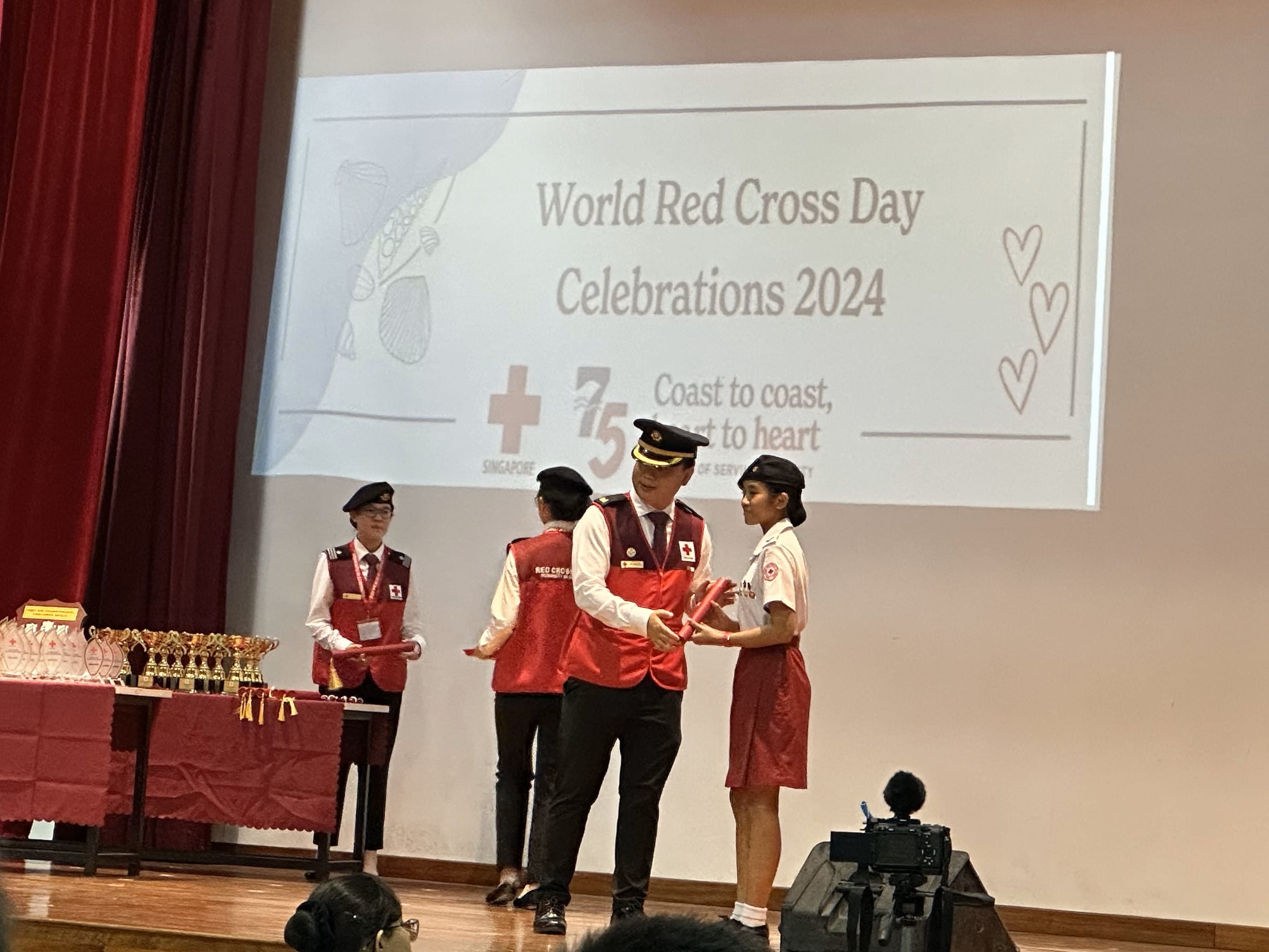People in Red Cross uniforms on stage at World Red Cross Day Celebrations 2024, with trophies on a table nearby.