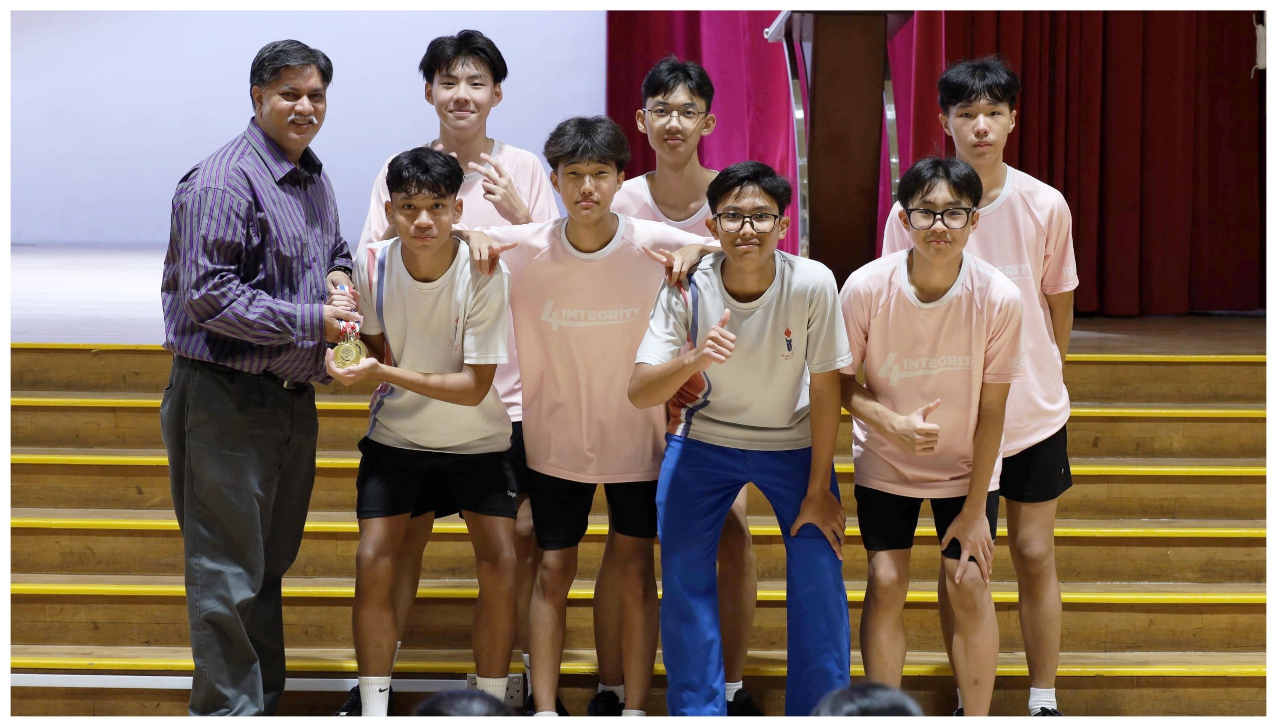 An adult man in a striped shirt holds a gold medal alongside six teenage boys in pink shirts on stage steps.