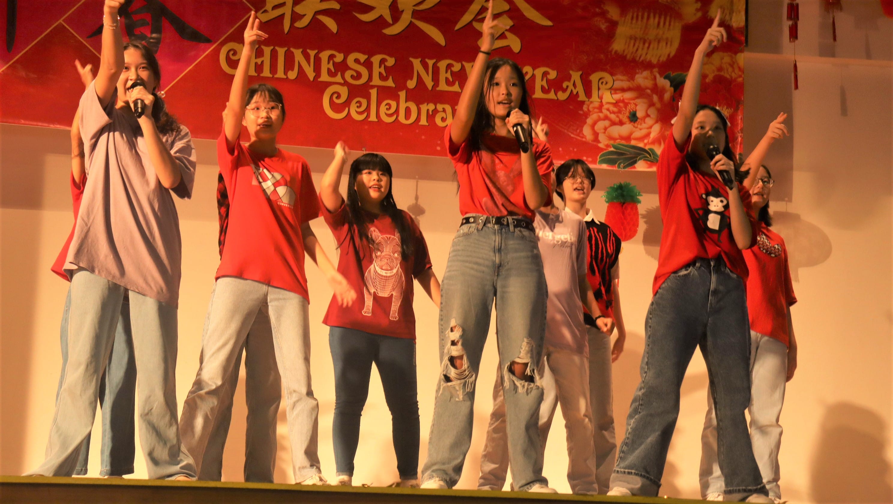 Young performers sing on stage at a Chinese New Year Celebration, several holding microphones and raising hands.