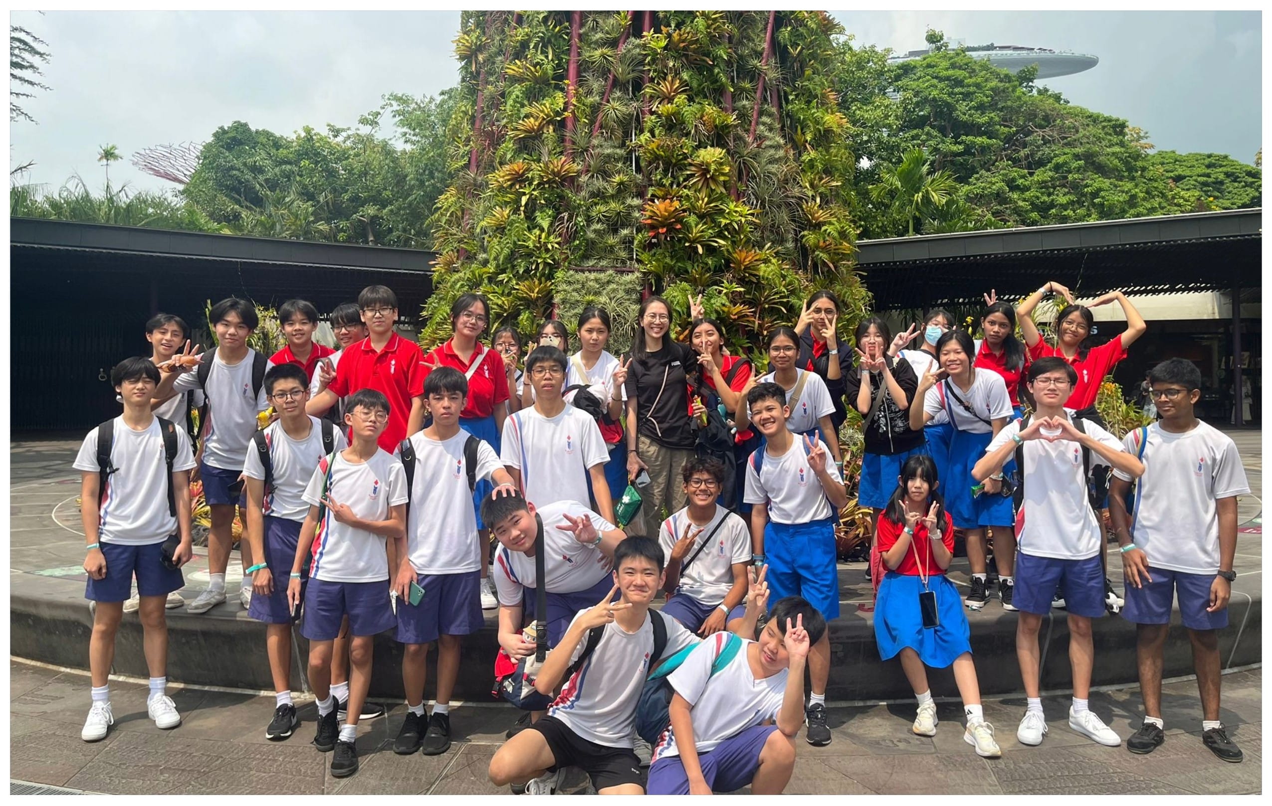 A large group of students in white and red uniforms pose with peace signs in front of a tall plant tower outdoors.