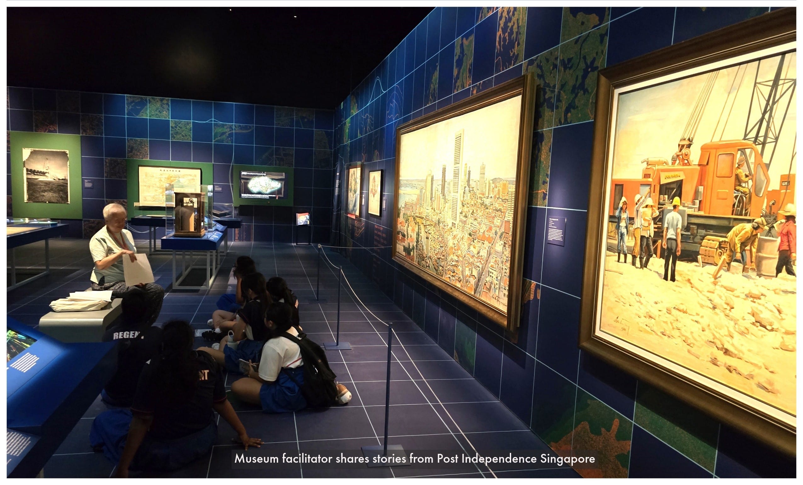 A museum facilitator speaks to students sitting on the floor beside large paintings of Singapore's cityscape and construction scenes.