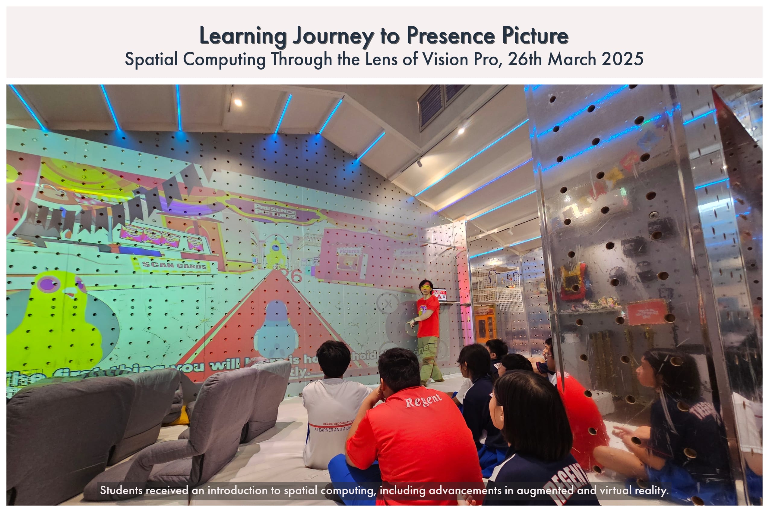 A presenter in a red shirt stands before students seated on beanbags in a colorful, tech-themed room with projected walls.