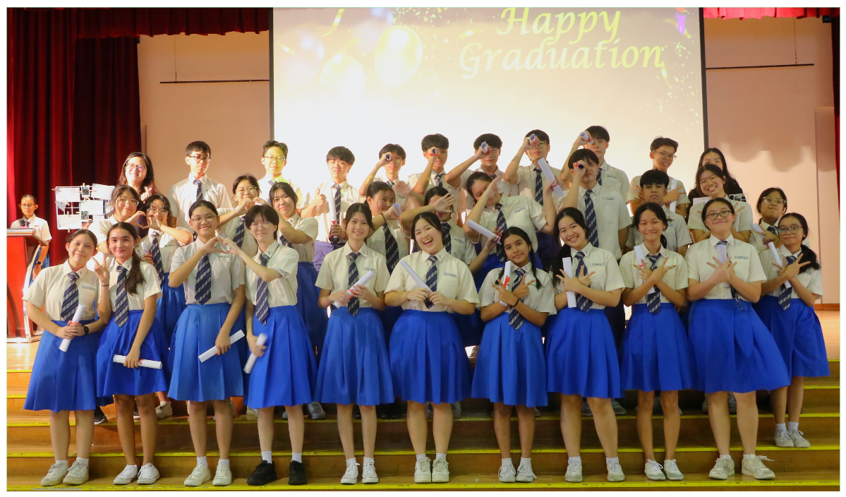 Students in white shirts and blue skirts pose on a stage in front of a screen reading Happy Graduation.