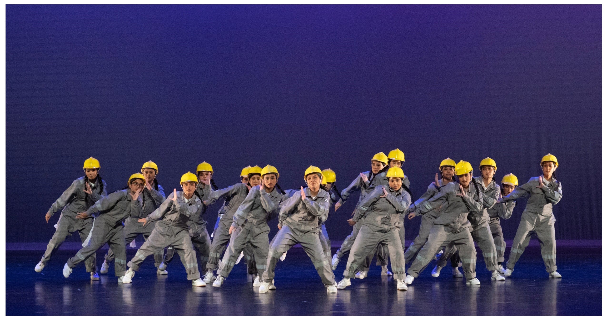 A large group of dancers in gray jumpsuits and yellow hard hats performing on a blue-lit stage.