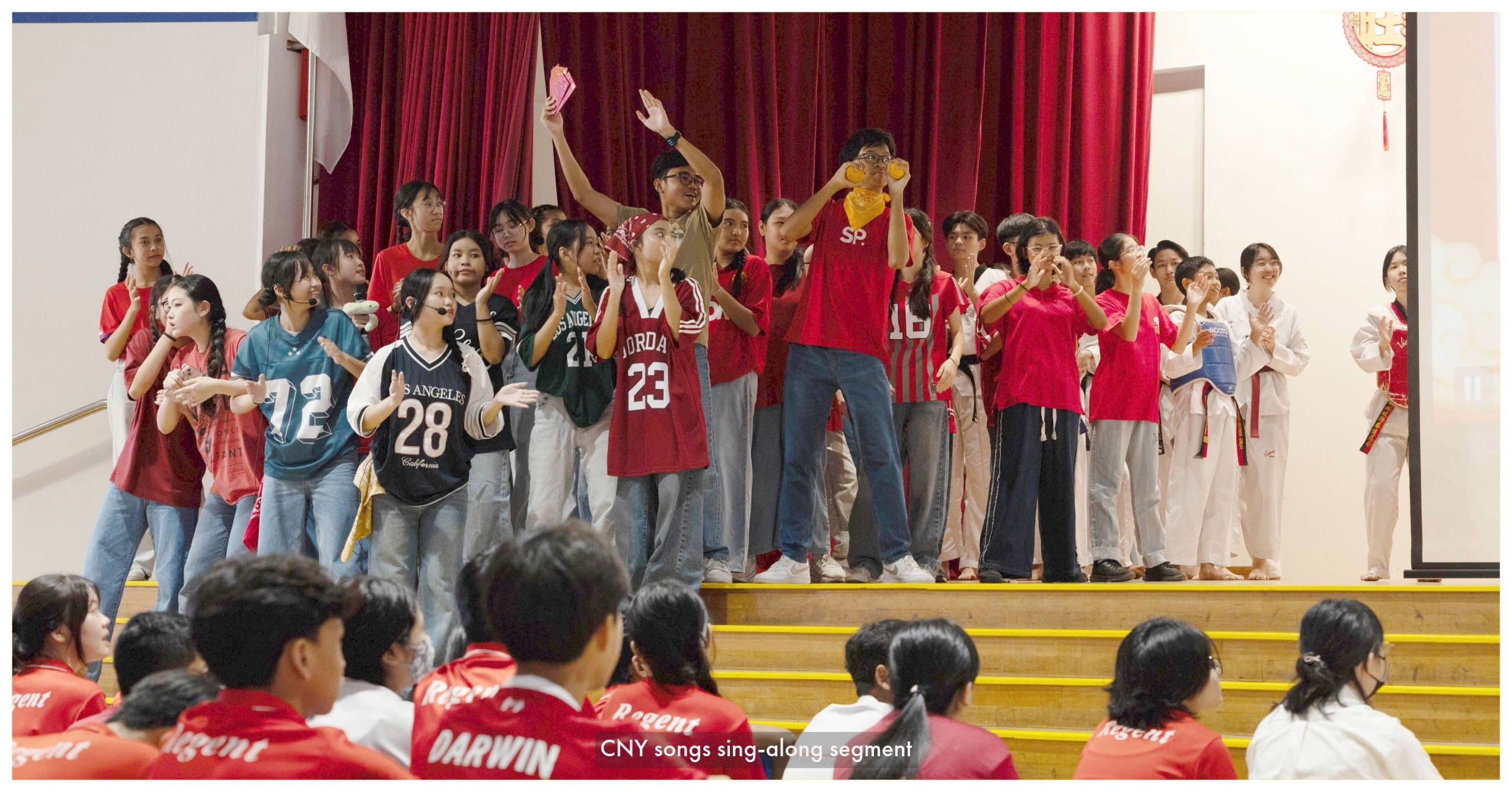 Students in red shirts and jerseys perform on a stage with red curtains during a CNY sing-along segment.