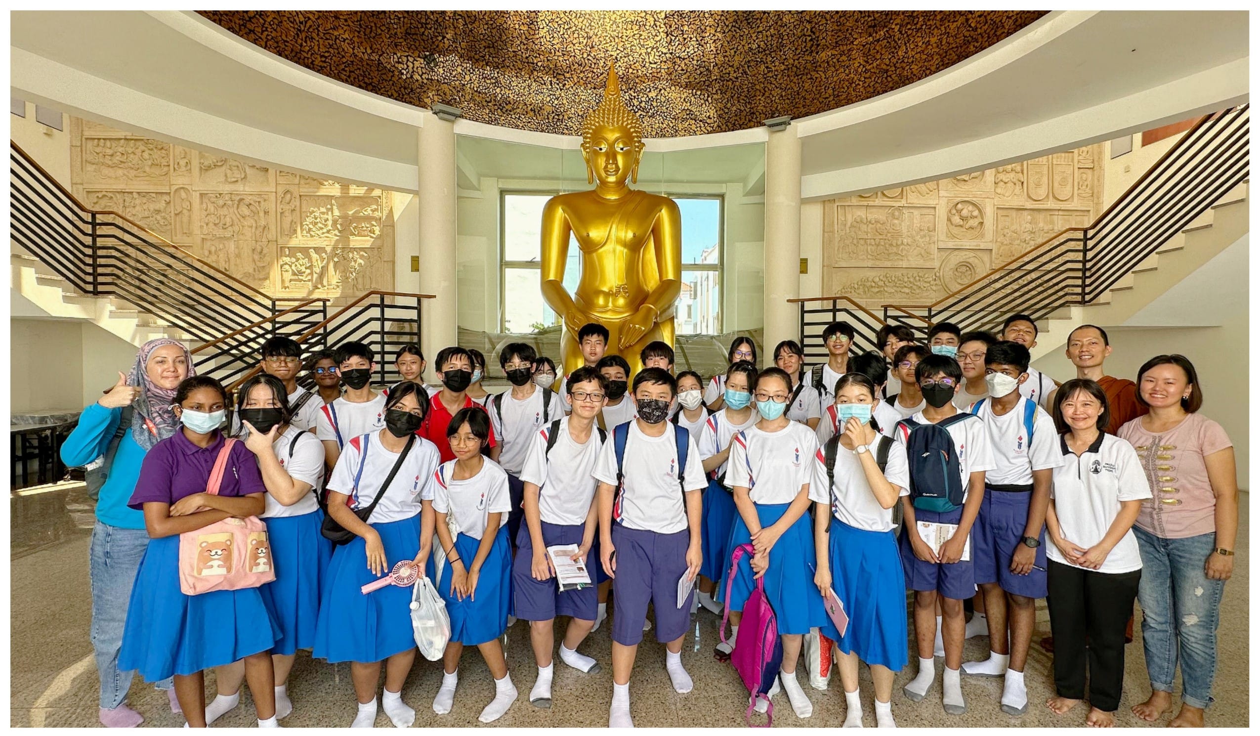 A group of students in white and blue uniforms pose in front of a large golden Buddha statue inside a temple hall.