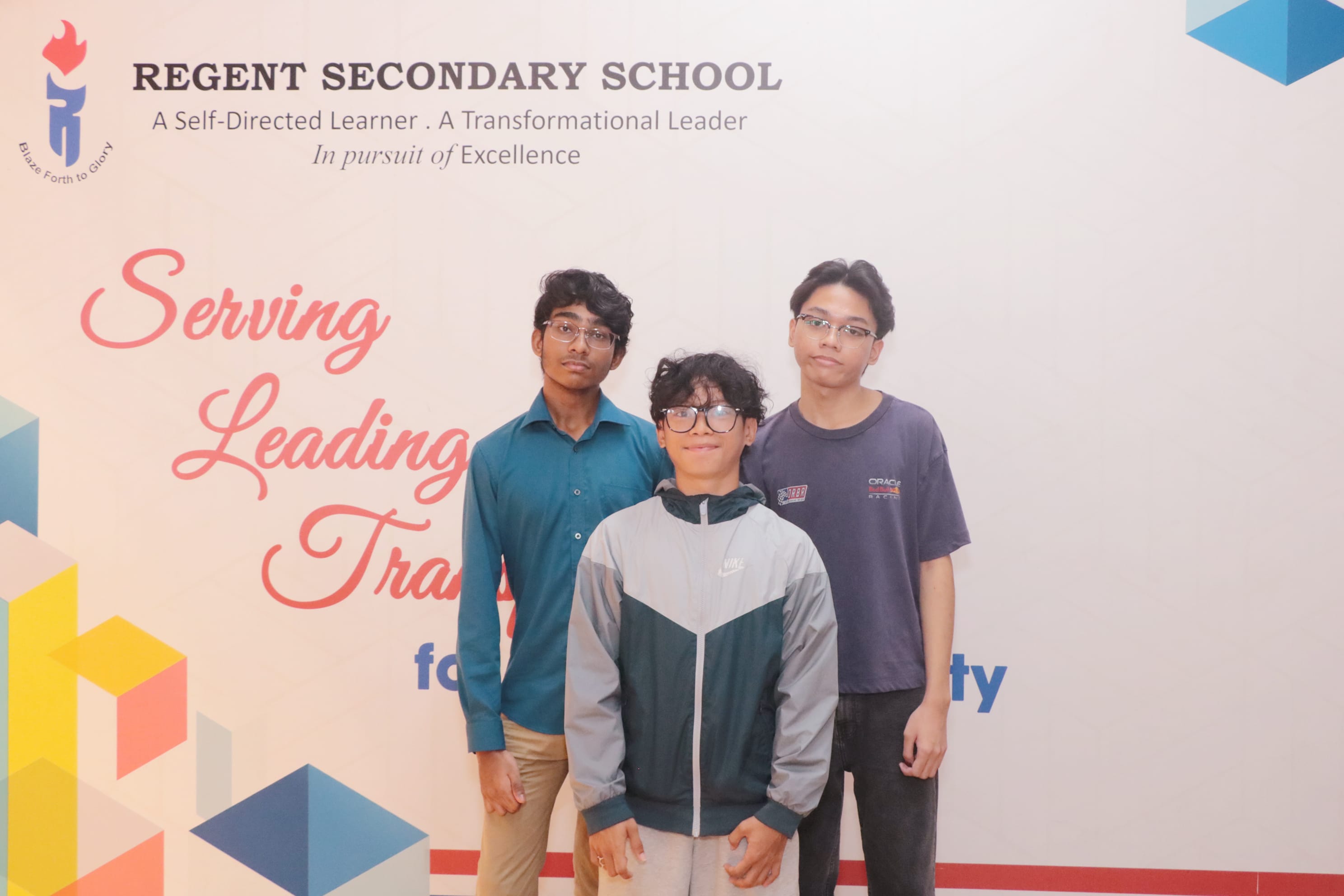 Three young students pose in front of a Regent Secondary School banner reading Serving, Leading, Transforming.