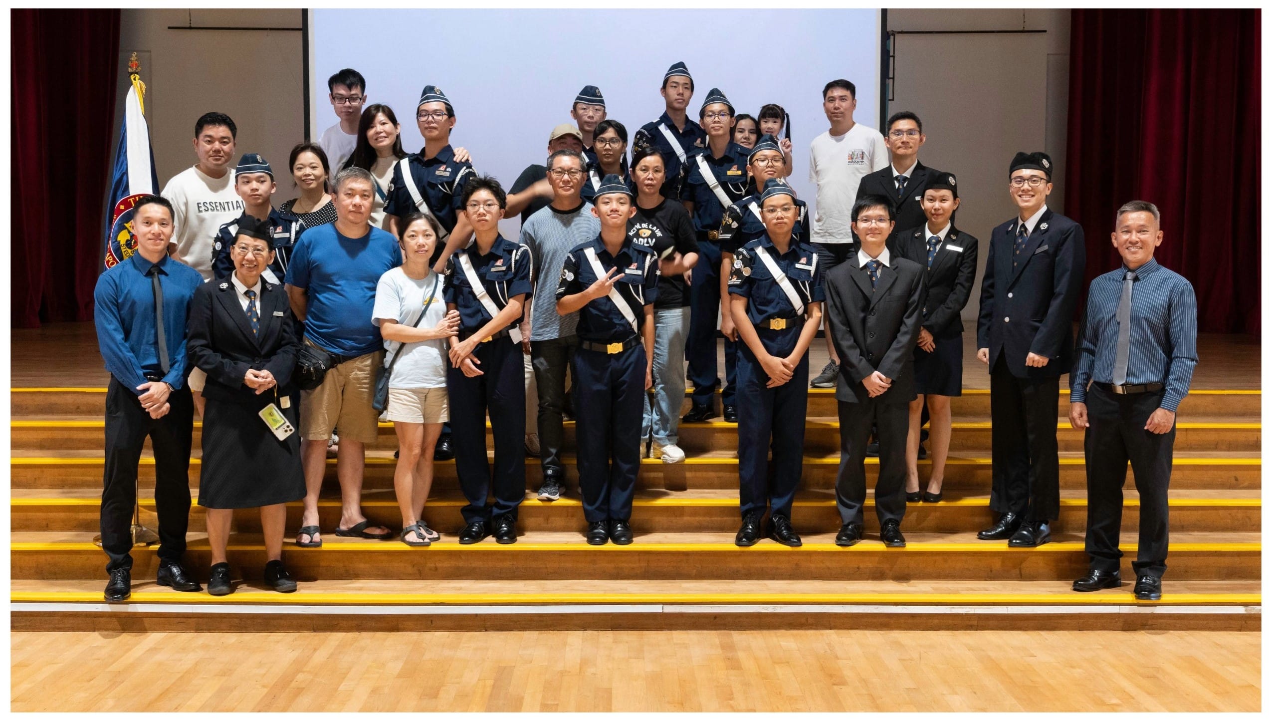 A large group of people posing on stage, several wearing navy blue uniforms with white sashes and caps.