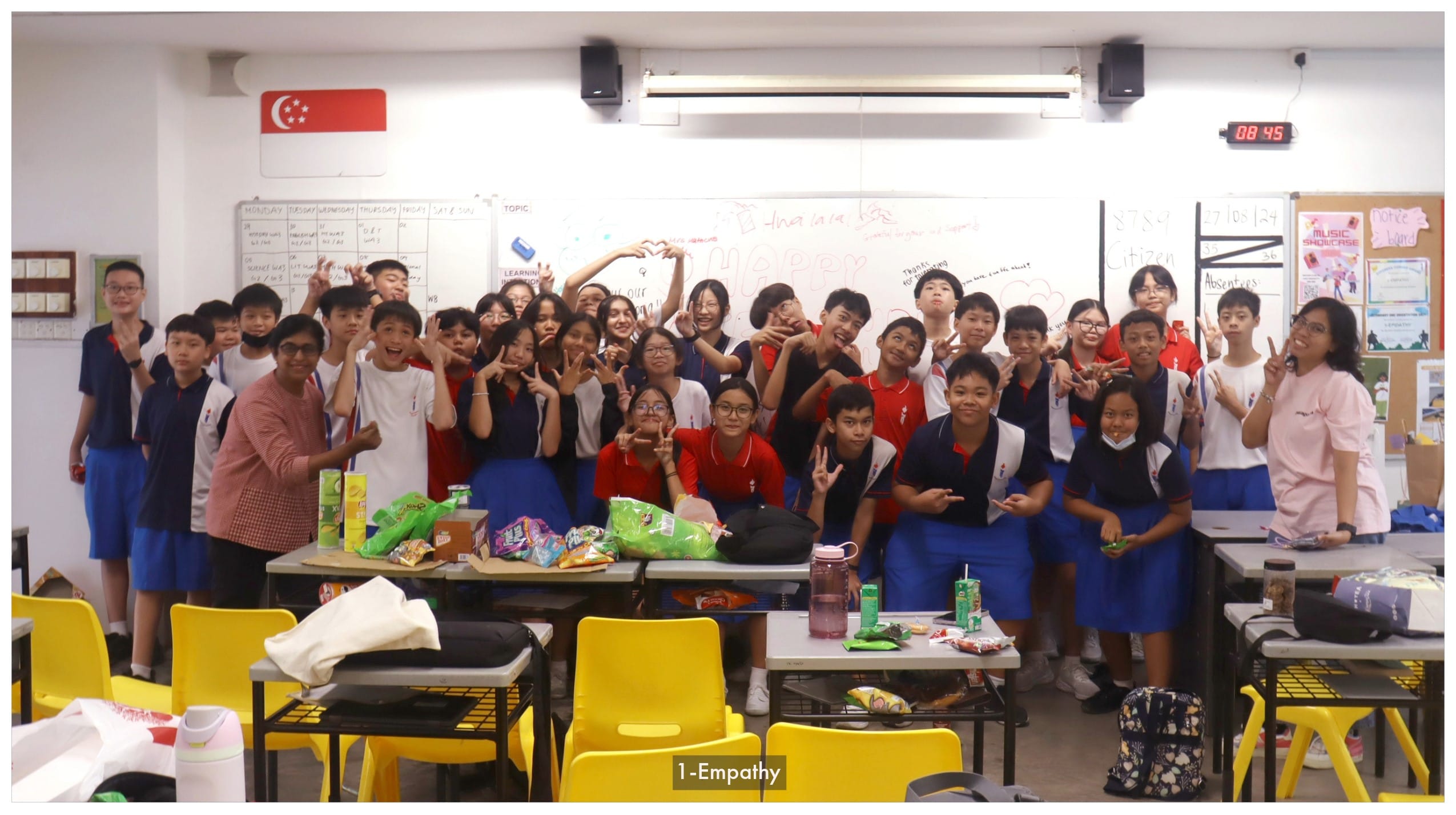 A large group of school students and a teacher pose cheerfully in a Singapore classroom with a whiteboard behind them.