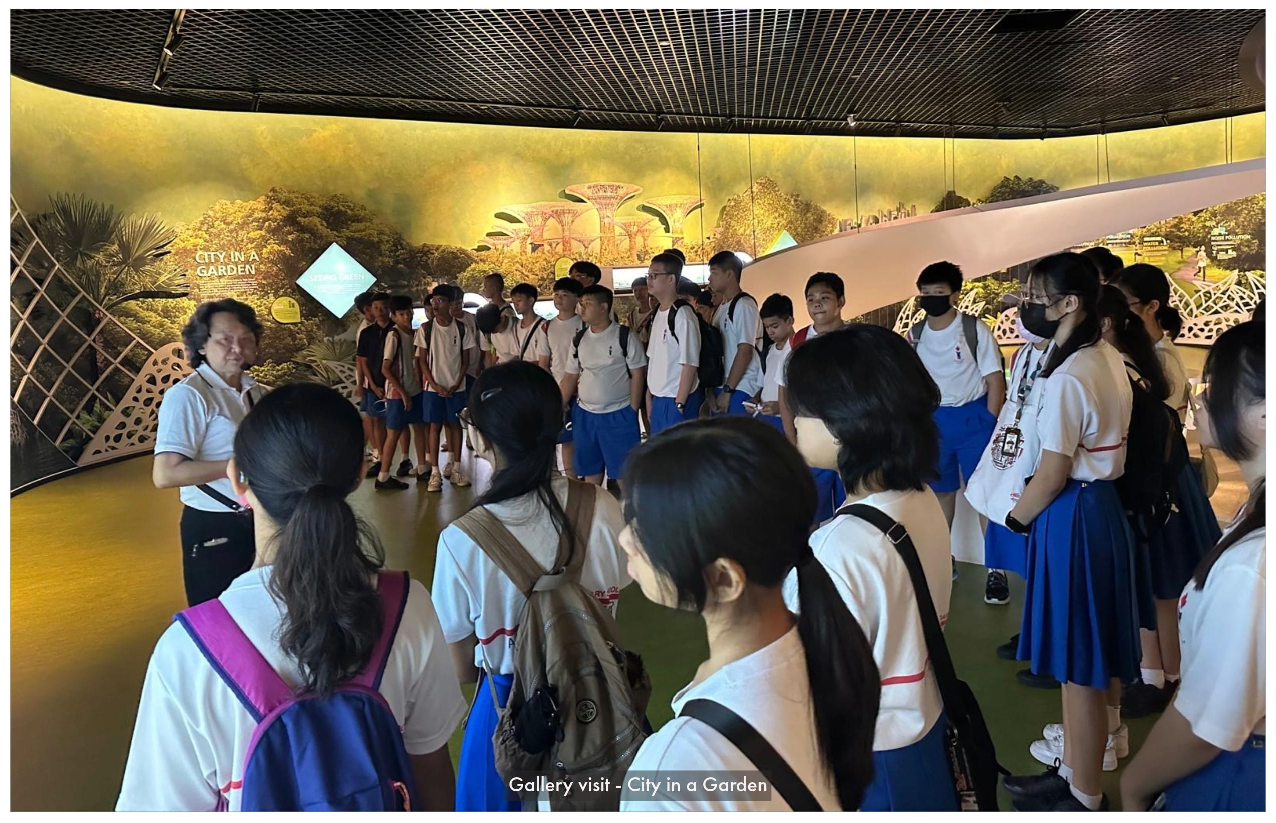 Students in white and blue uniforms listen to a guide in a City in a Garden exhibition room.