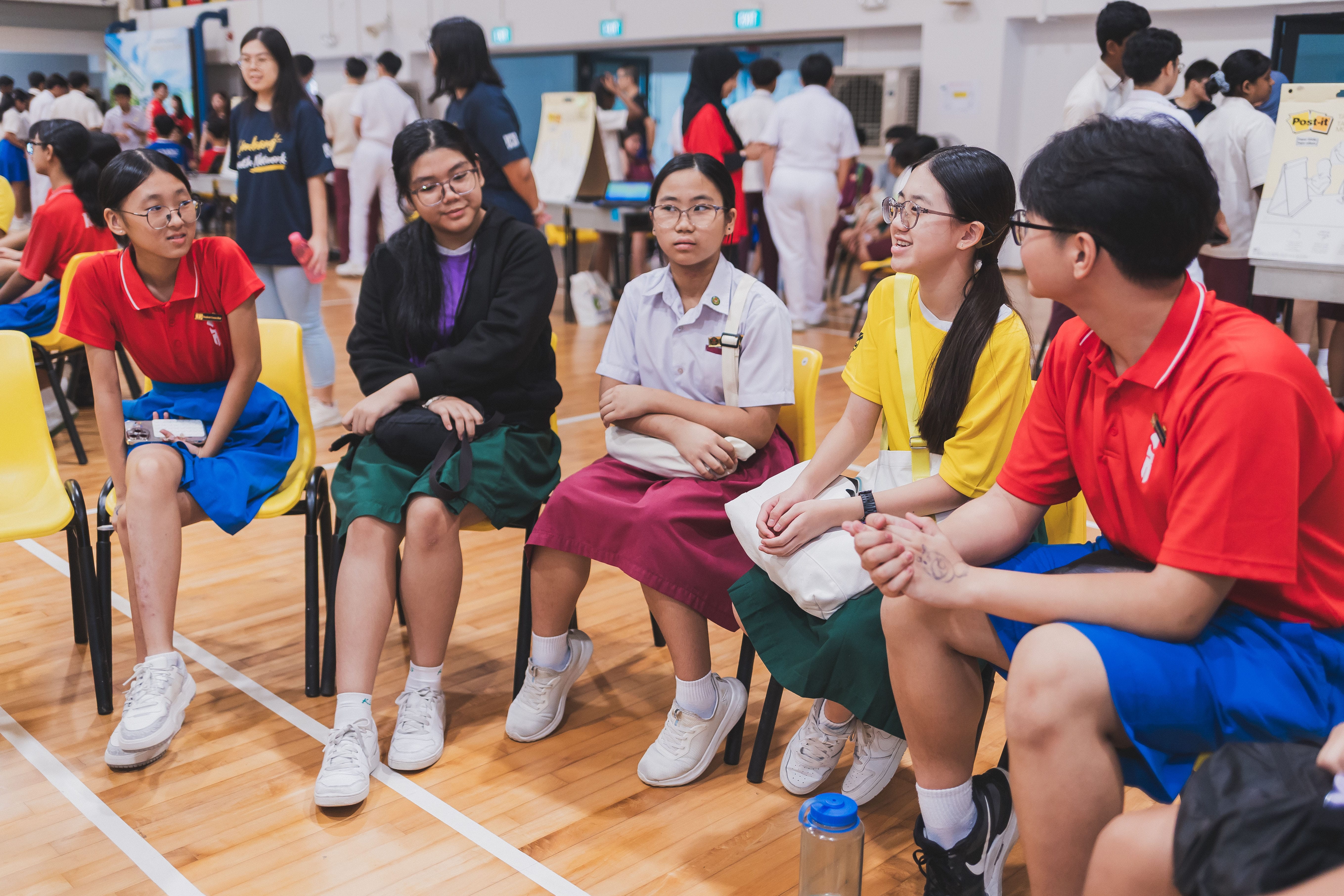 Five students in colorful school uniforms sit in a circle on yellow chairs in a busy school hall.