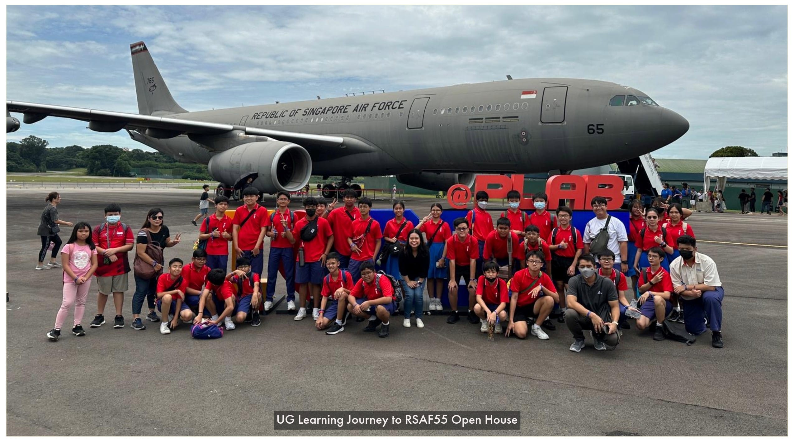 A group of students in red shirts pose in front of a Republic of Singapore Air Force aircraft on a tarmac.