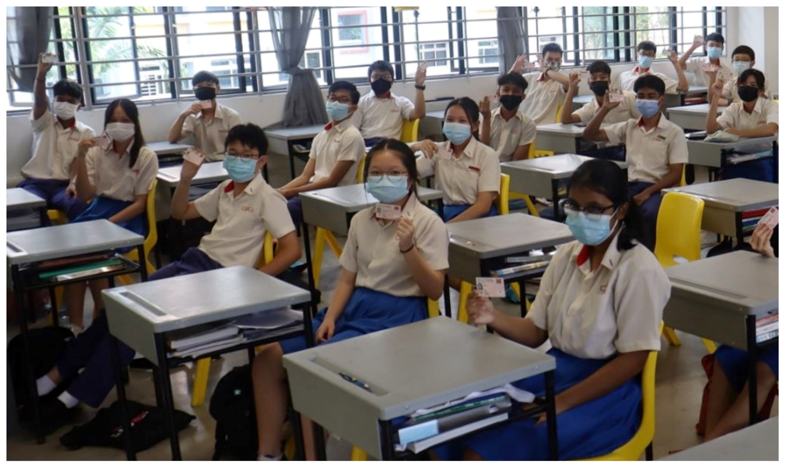Students in school uniforms and face masks sit at desks in a classroom, each holding up a small card.