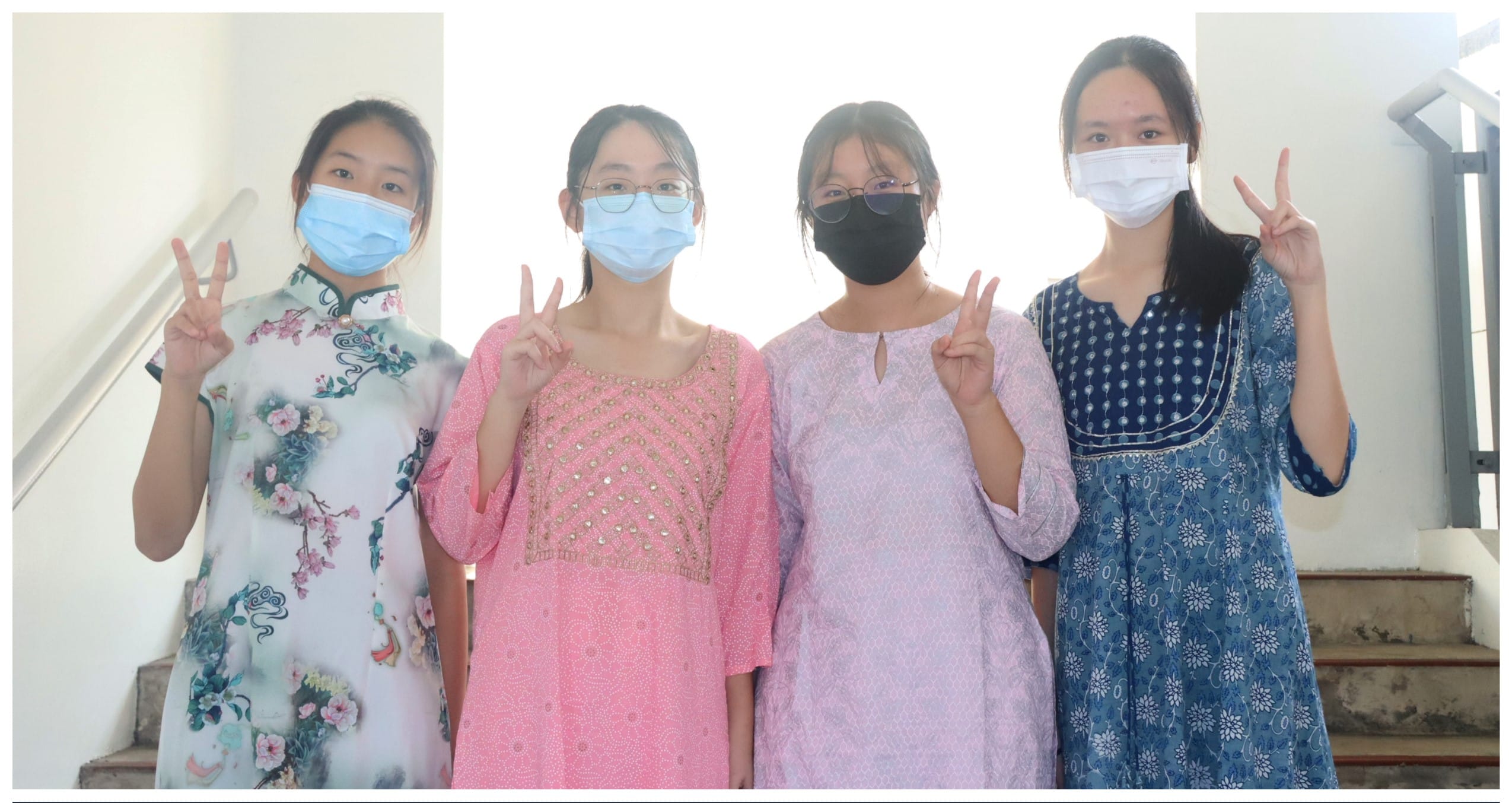 Four young women wearing face masks pose together on stairs, each making a peace sign with their hand.