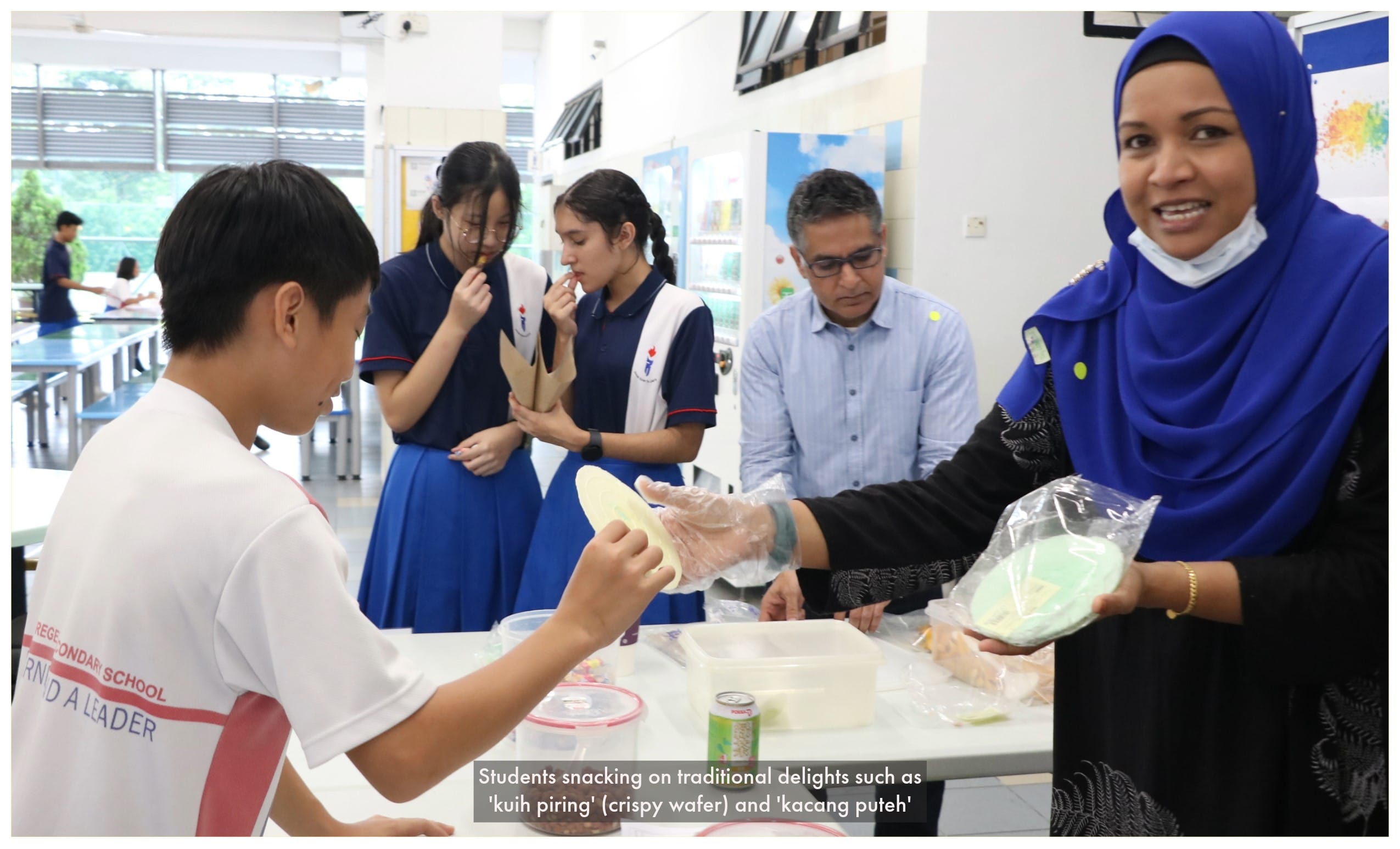 Students and adults gather around a table sampling traditional Malay snacks like kuih piring in a school canteen.
