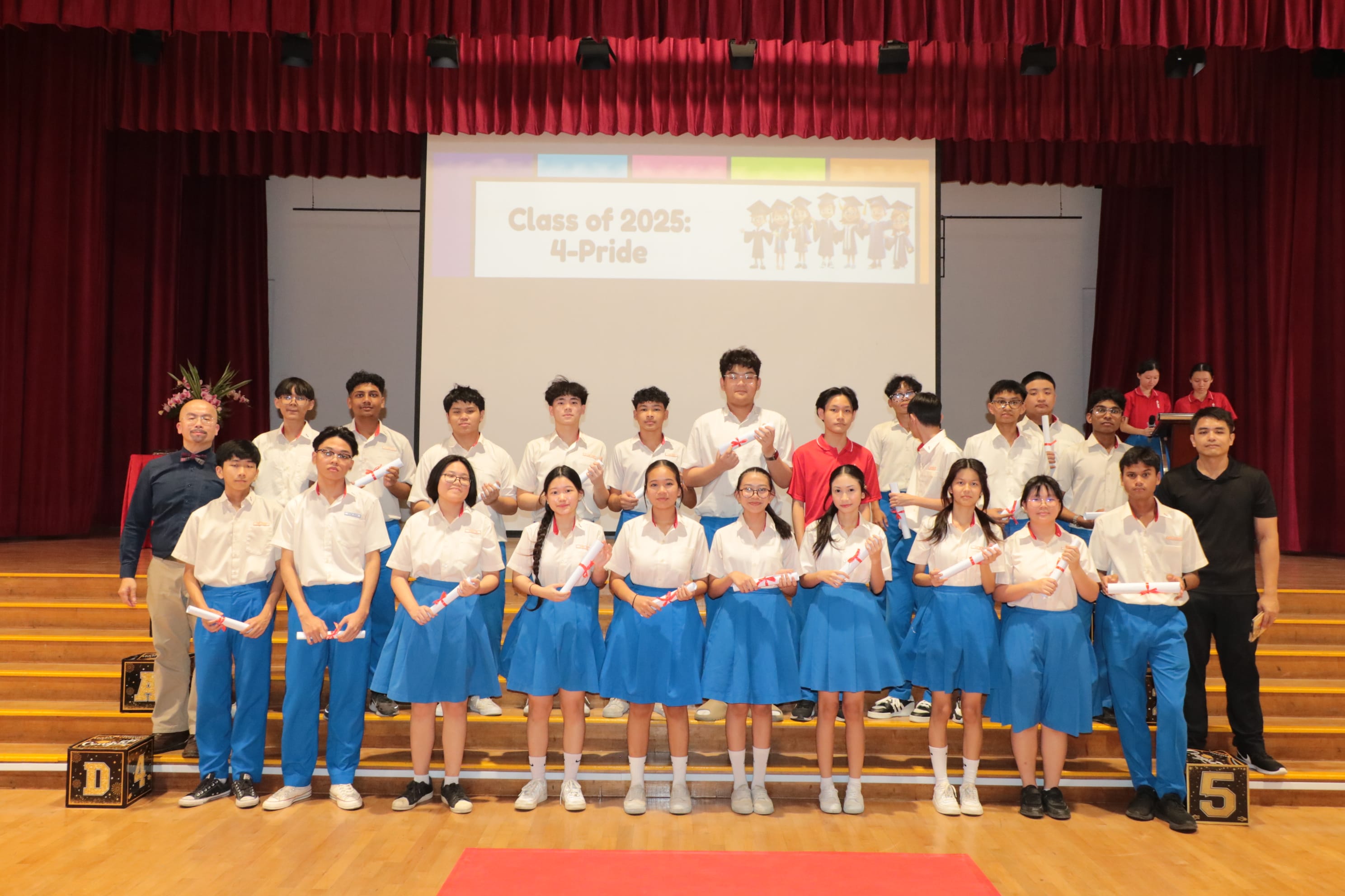 Students in white and blue uniforms hold scrolls on stage before a screen reading Class of 2025: 4-Pride.