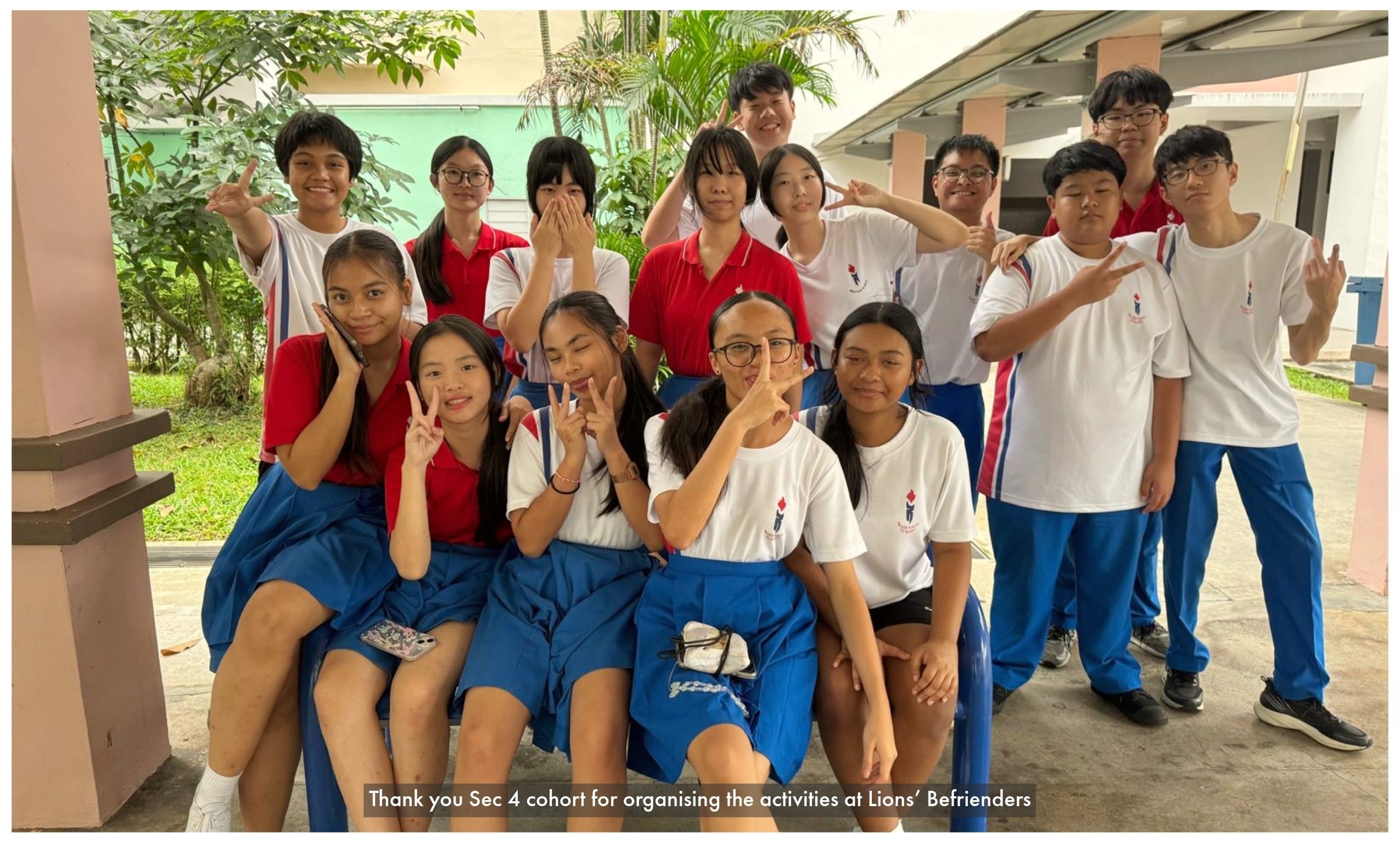 A group of teenage students in red and white uniforms pose and smile outdoors, making hand gestures for a group photo.