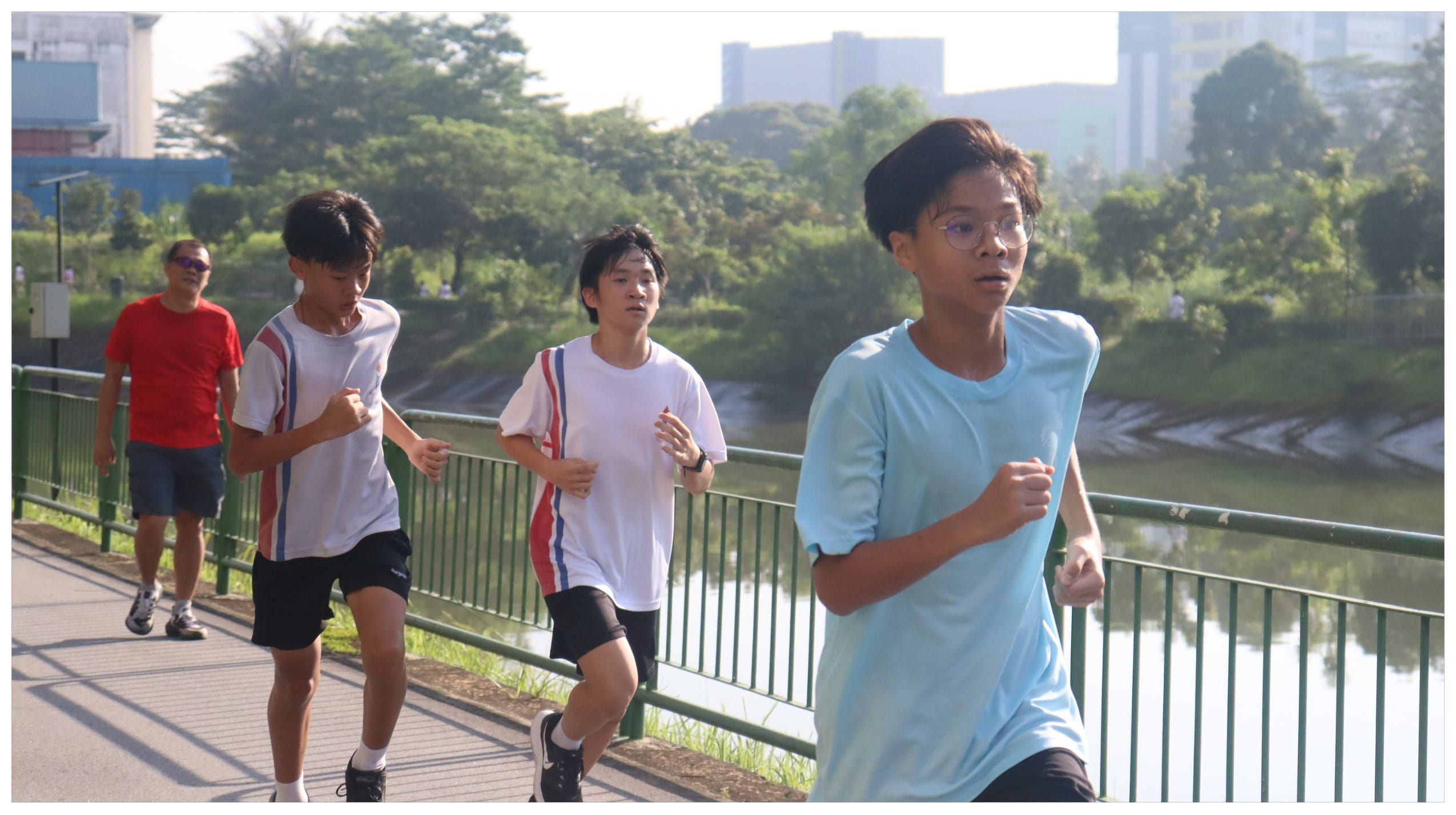 Three teenagers jogging on a riverside path with an adult walking behind them, trees and buildings in the background.