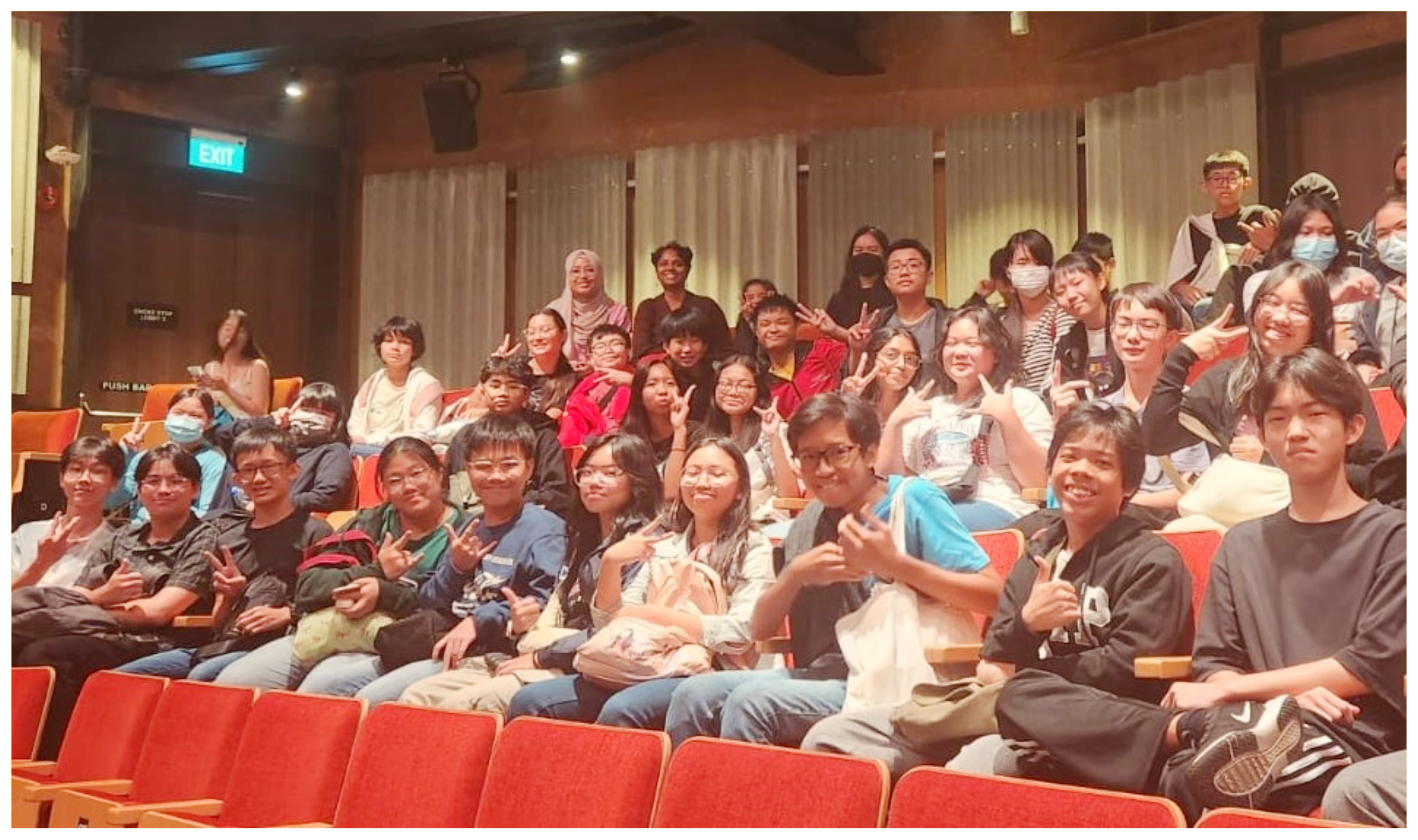 Large group of students seated in an auditorium with red seats, posing and making peace signs.