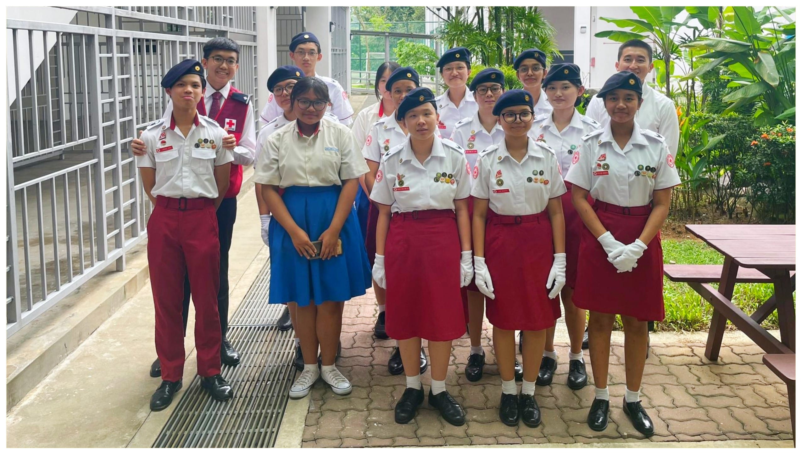 A group of students in Red Cross uniforms with navy berets pose together outdoors on a paved courtyard.