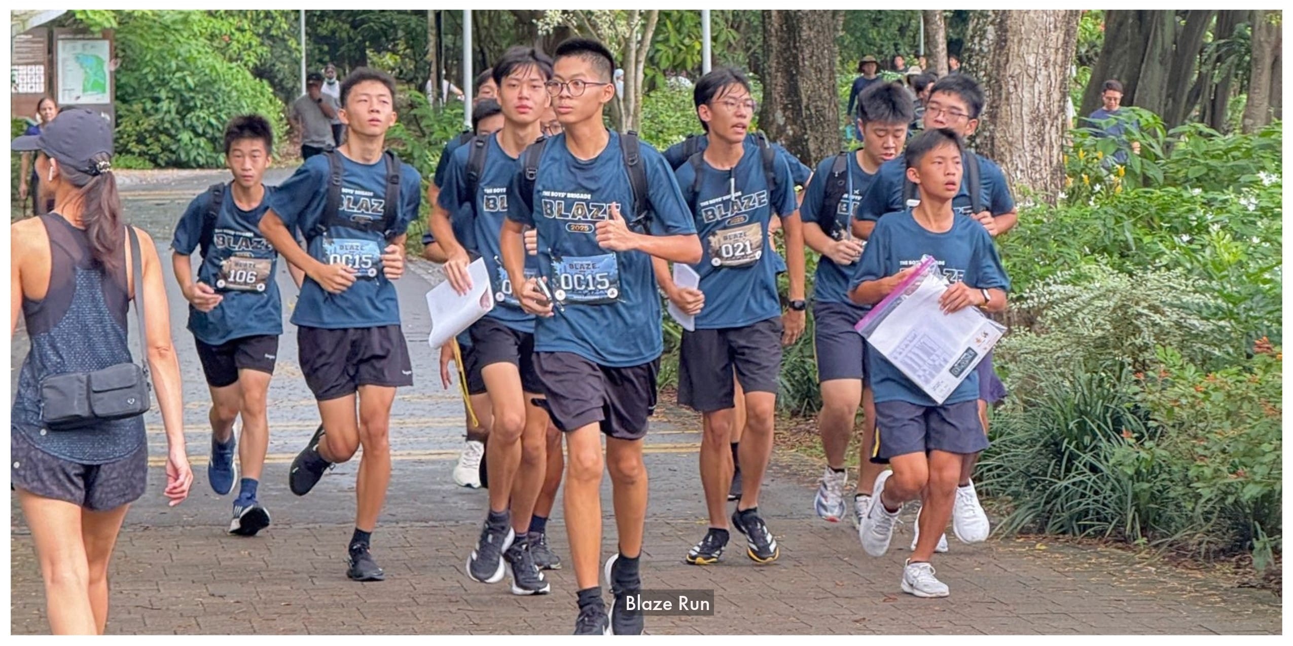A group of young male runners in matching blue Blaze shirts and race bibs jogging along a tree-lined path.