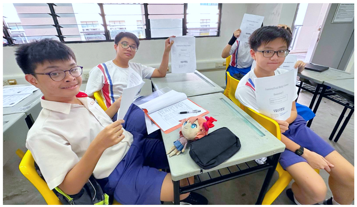 Three students in school uniforms sit at desks holding up papers, with a small doll and pencil case on the desk.
