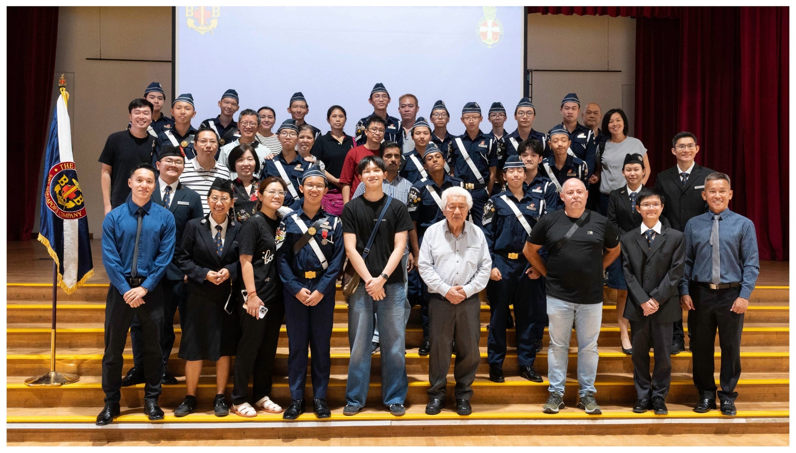 A large group of youth in navy uniforms with sashes pose with adults on auditorium steps beside a ceremonial flag.
