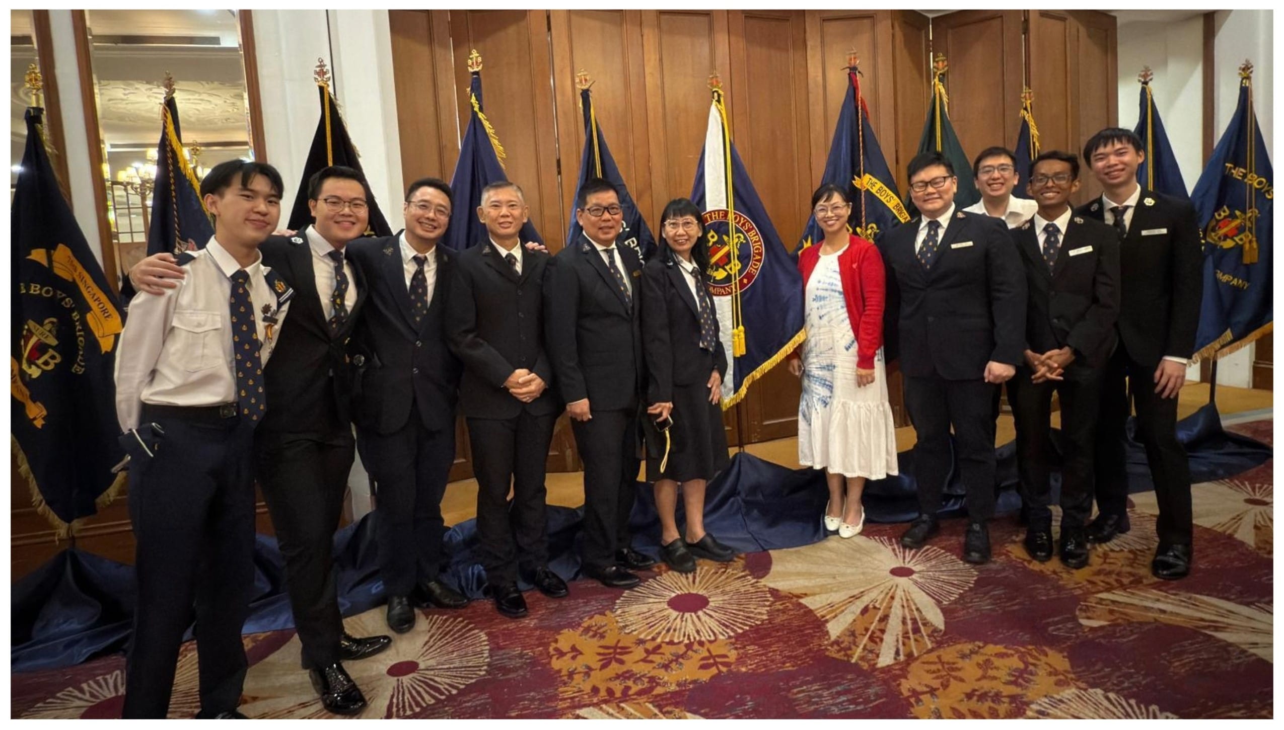 Group of about twelve people in formal attire posing together in front of several Boys Brigade flags indoors.