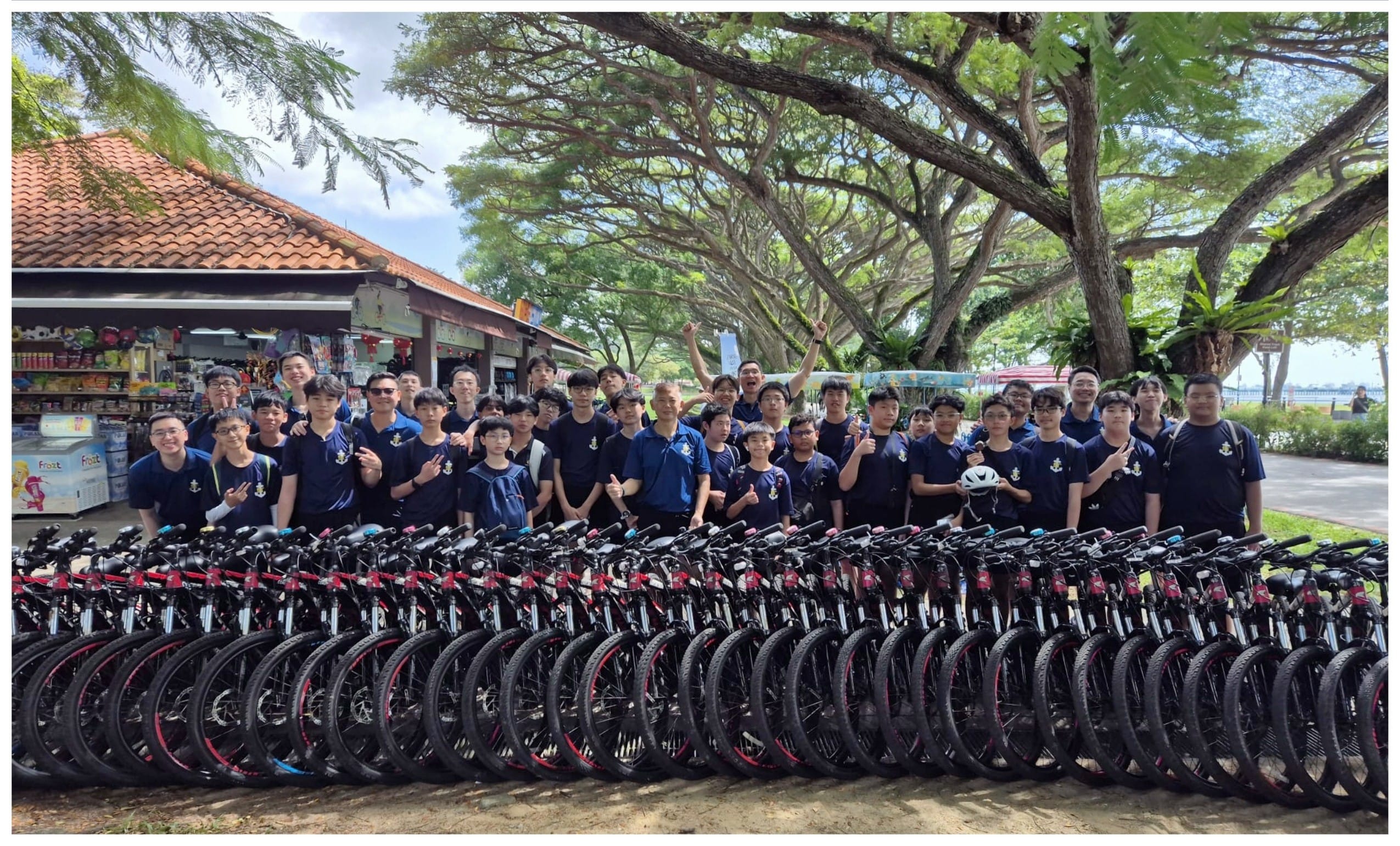 A large group of students in blue uniforms posing behind a row of bicycles outdoors near trees and a small shop.