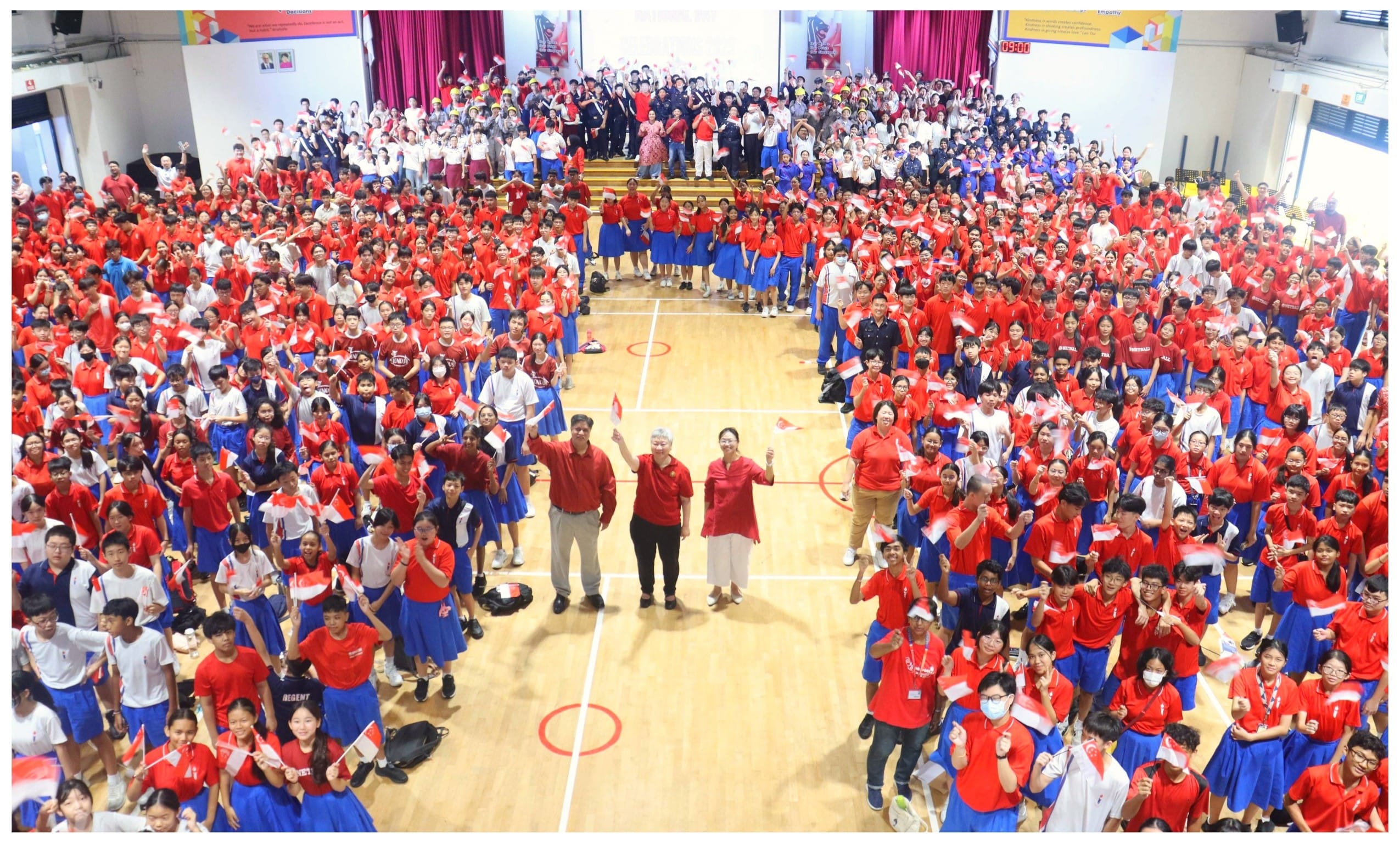 Large crowd of students in red and blue uniforms waving Singapore flags in a school hall.