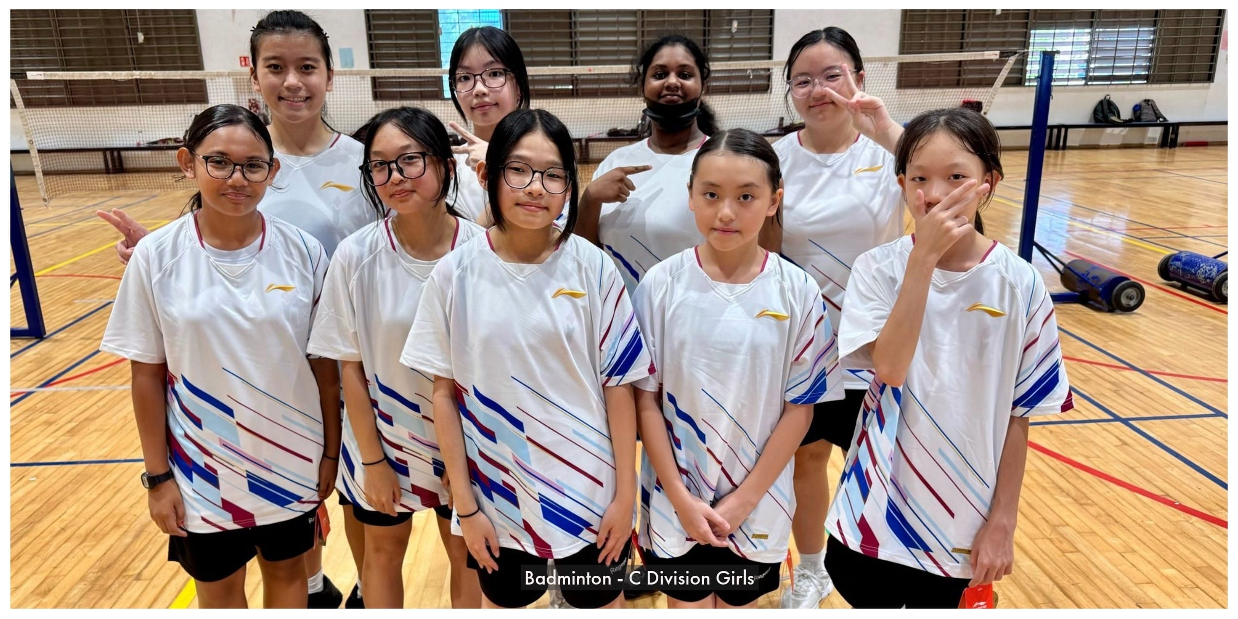 Eight girls in matching white sports jerseys pose together on an indoor badminton court.