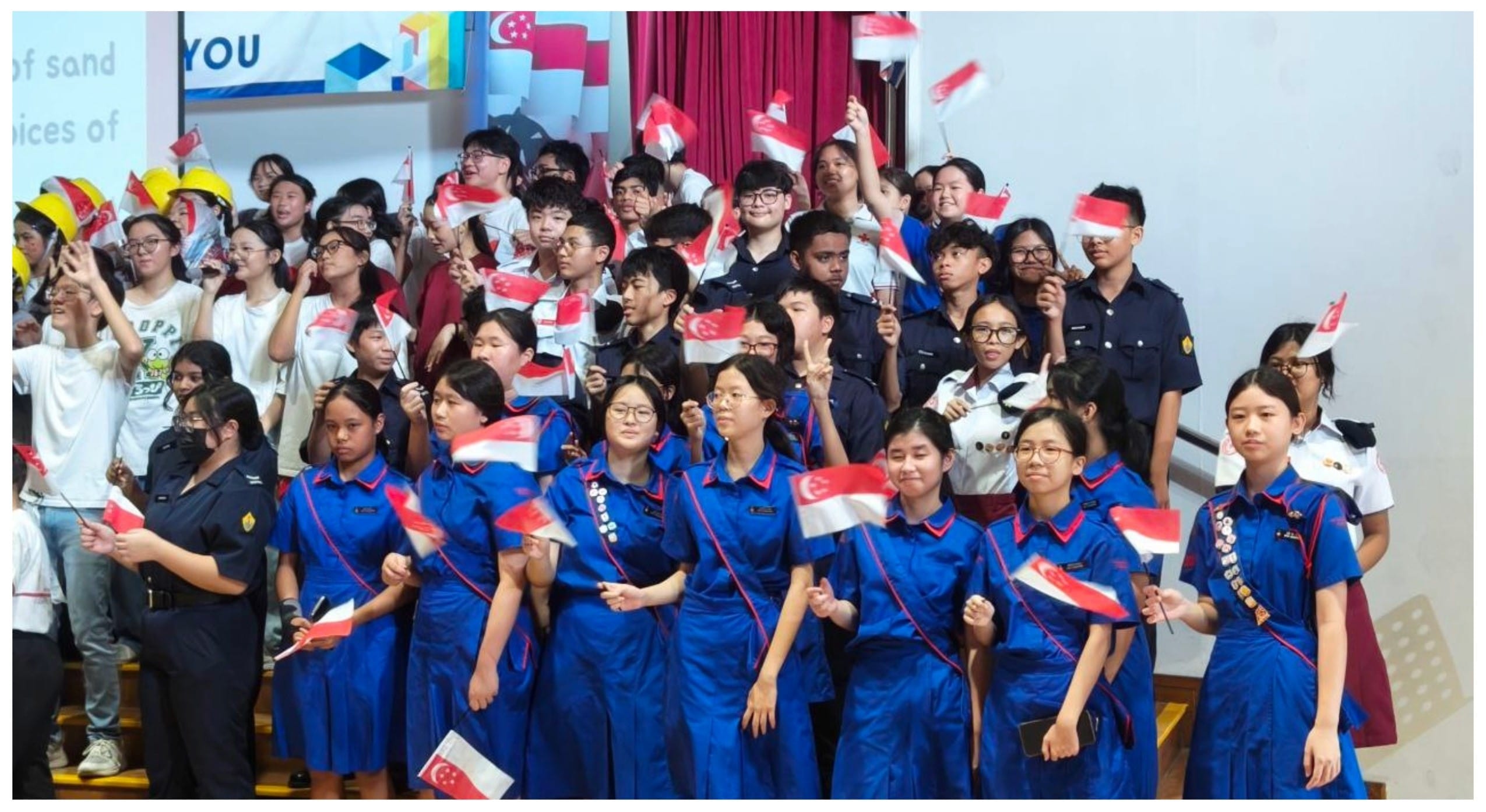 Students in uniforms wave Singapore flags during a school celebration event indoors.