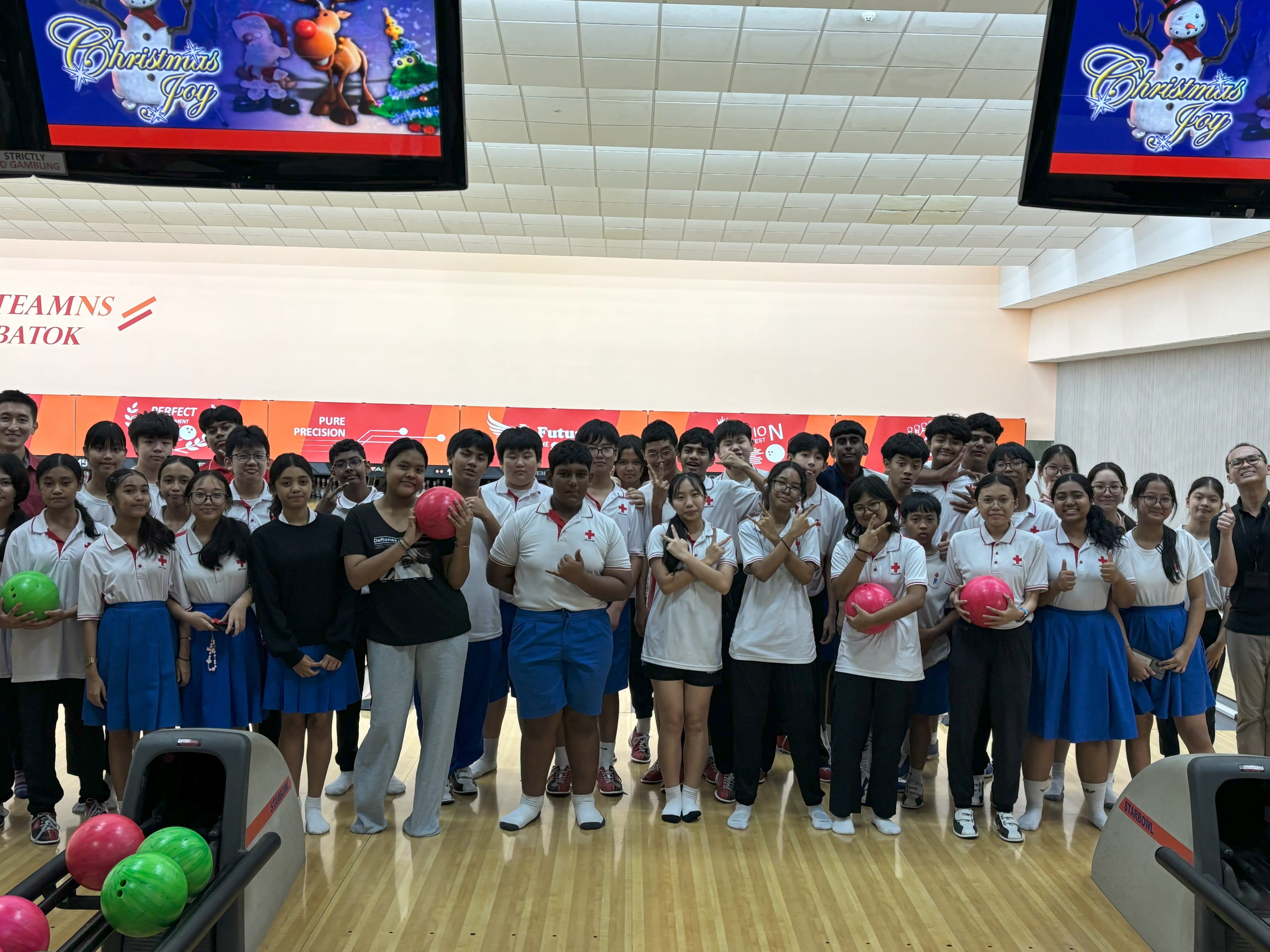 A large group of students in white uniforms pose together at a bowling alley with Christmas Joy screens above.