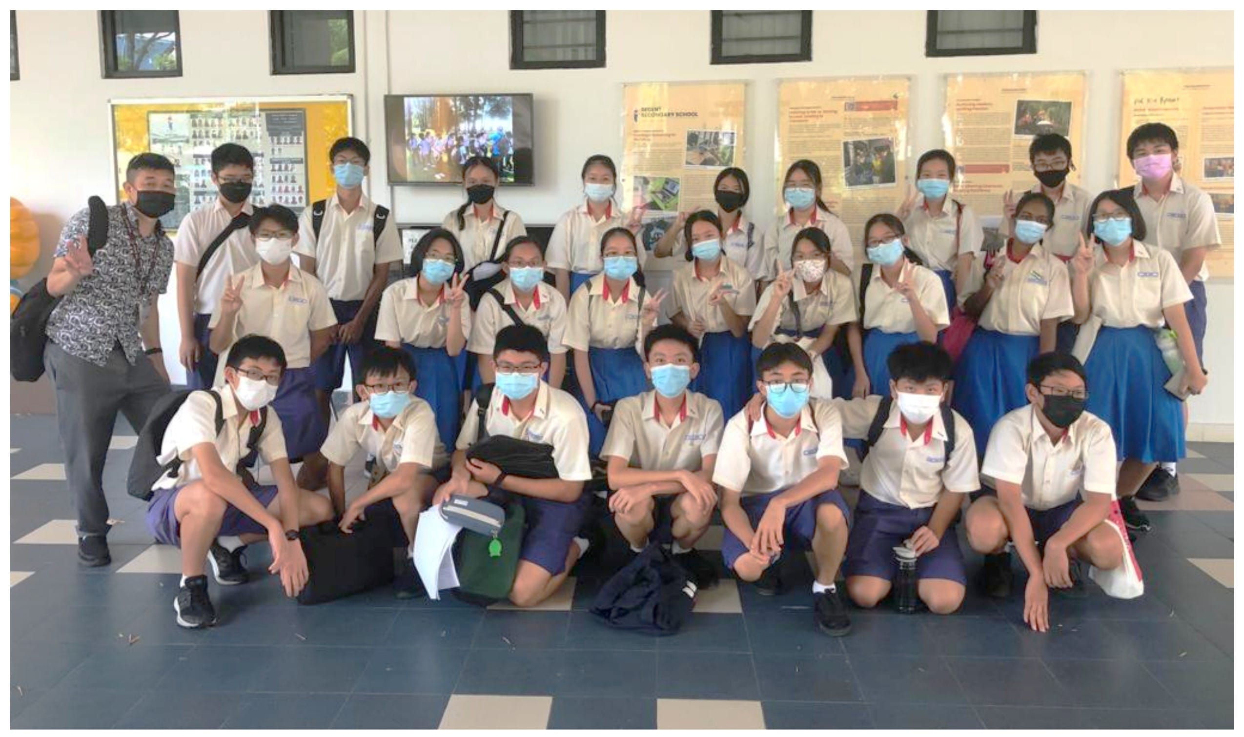 A large group of uniformed students and a teacher wearing face masks pose together indoors for a group photo.