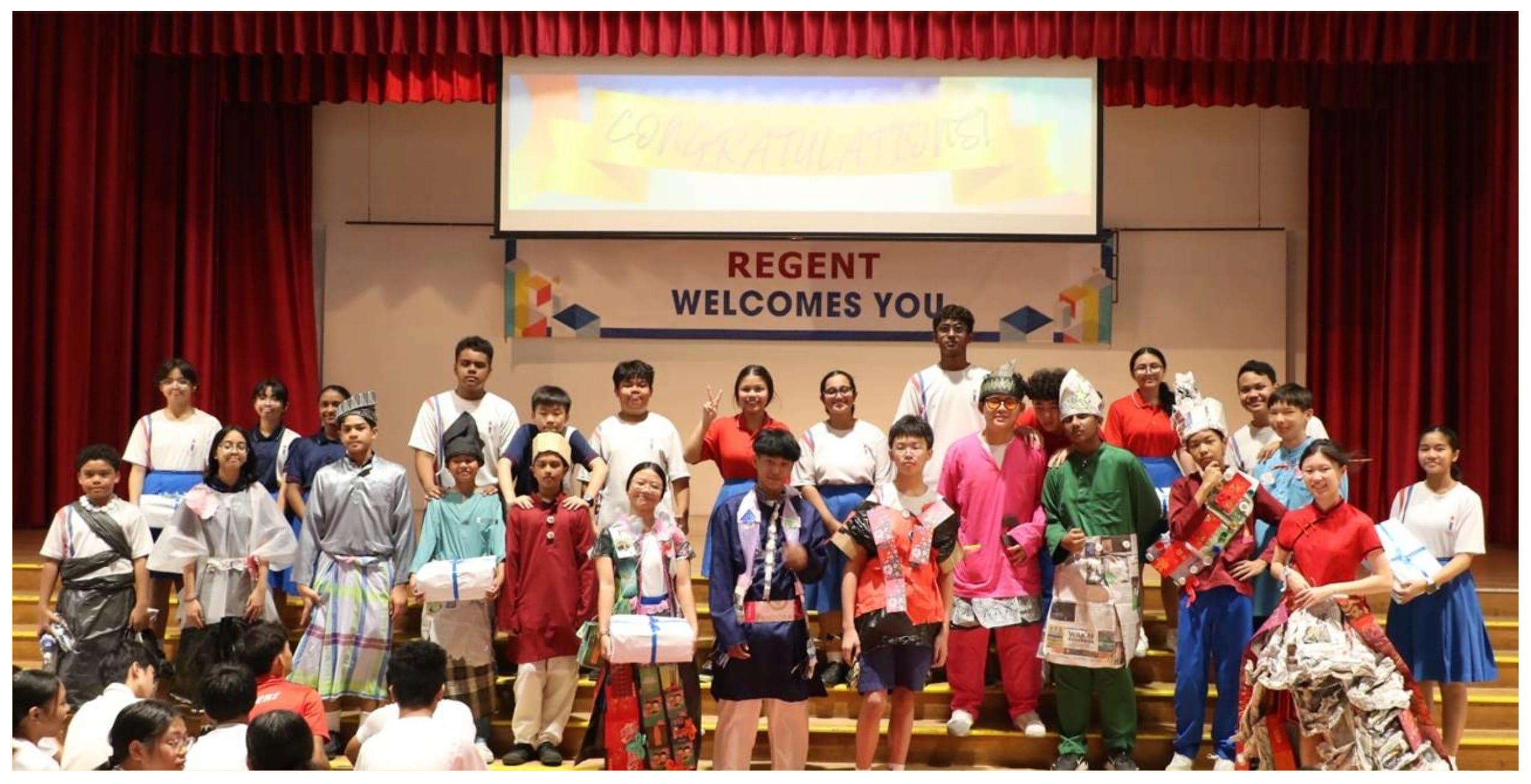 A large group of students in colorful costumes pose on a stage with a banner reading Regent Welcomes You.