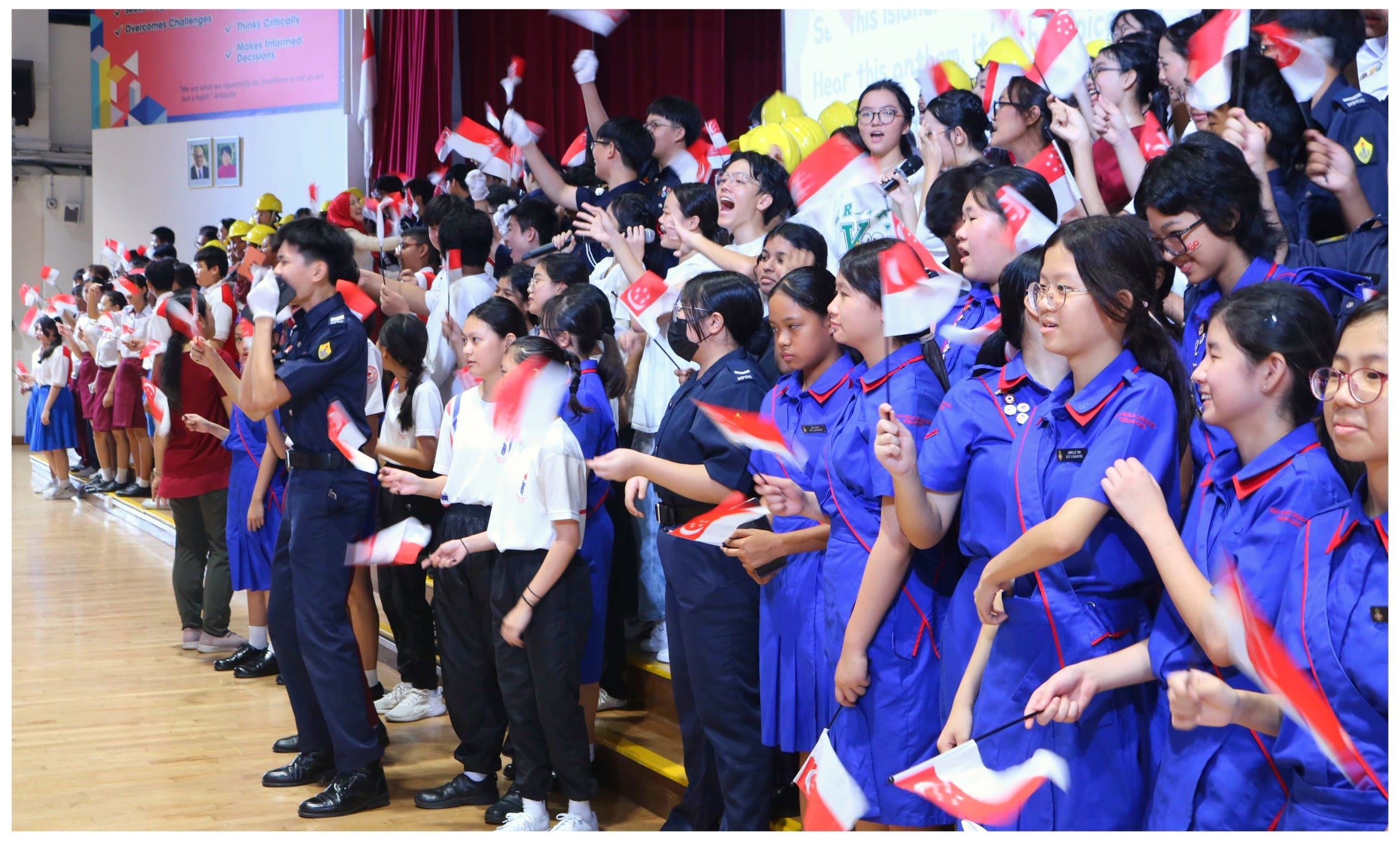 Students in uniforms wave Singapore flags and cheer enthusiastically inside a school hall.