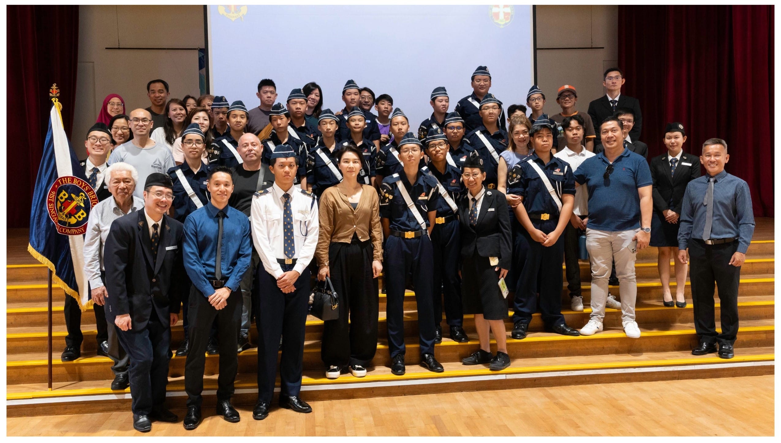 Large group of youth and adults in Boys Brigade uniforms posing on stage steps with a brigade flag nearby.