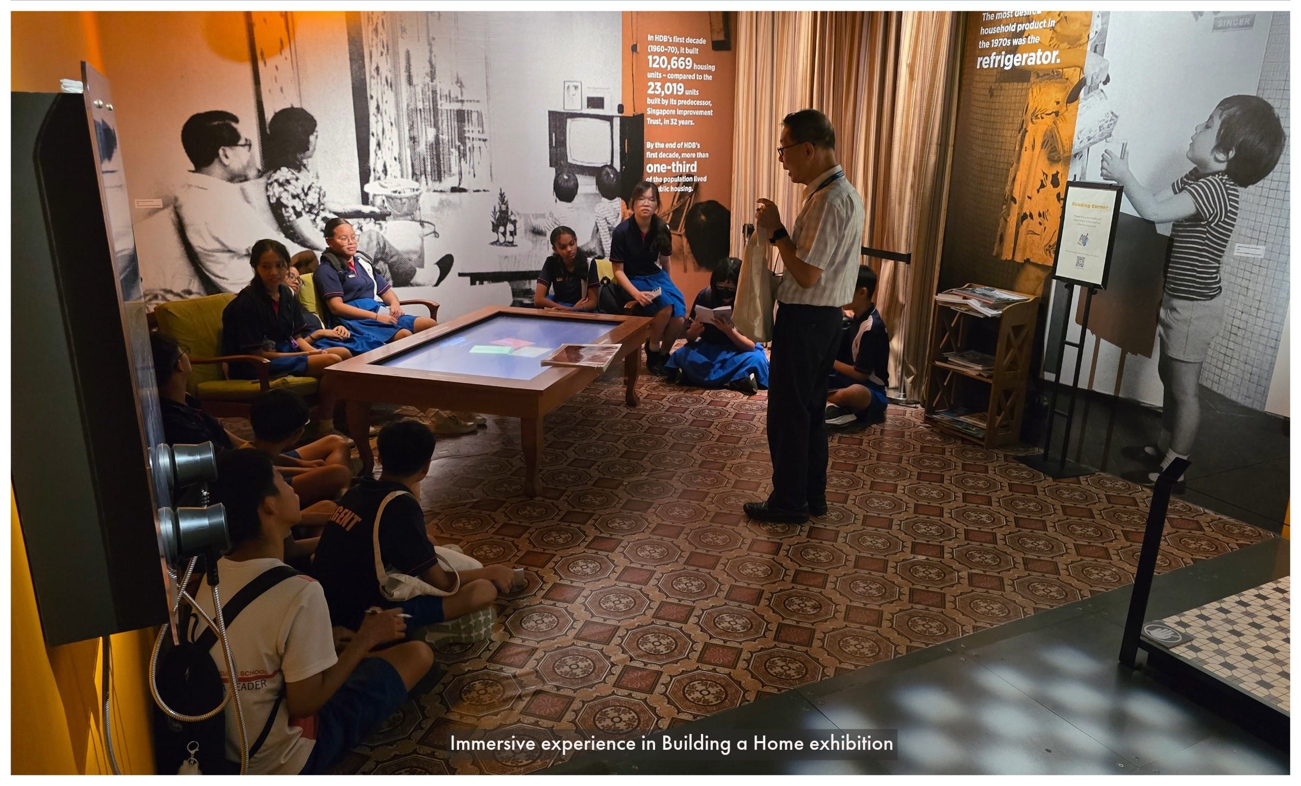 A guide speaks to students seated around a touch table in a museum exhibition room with historical photo murals.