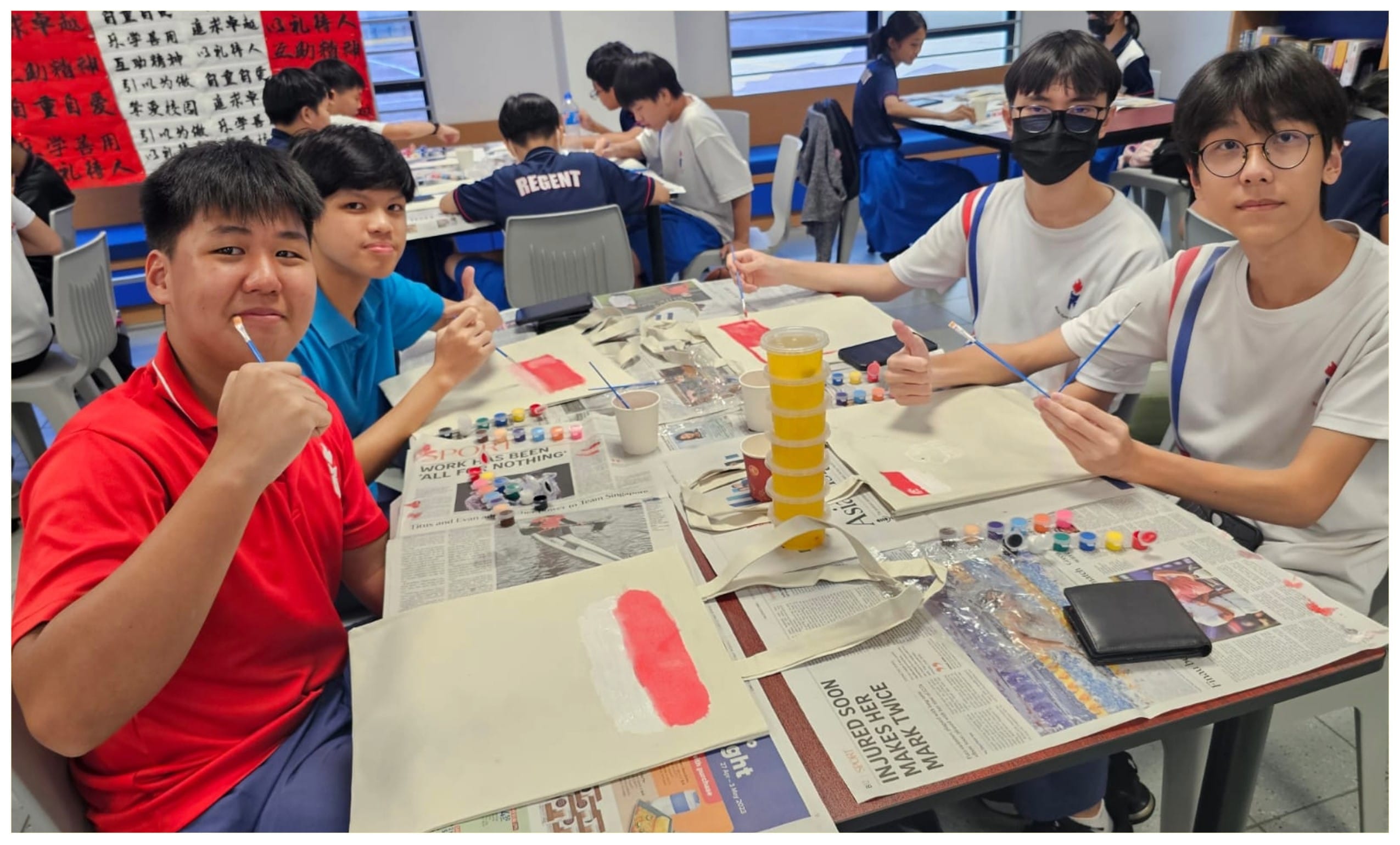 Four teenage students sit around a table covered in newspaper, painting with small brushes and acrylic paints in a classroom.