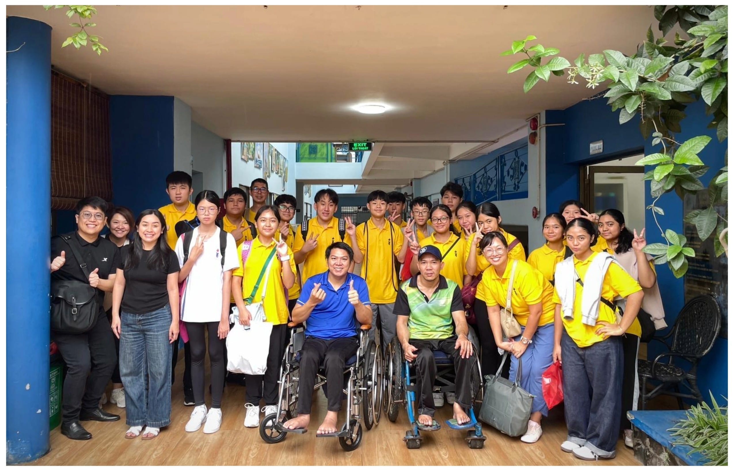 A large group in yellow uniforms poses indoors with two men in wheelchairs at the front, smiling and giving thumbs up.