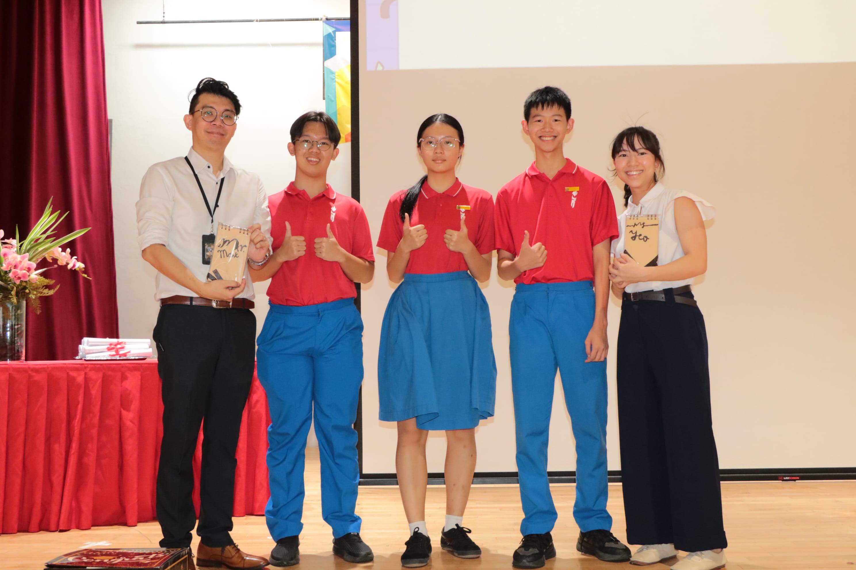 Five people pose on a stage, three students in red and blue uniforms giving thumbs up, flanked by two adults.