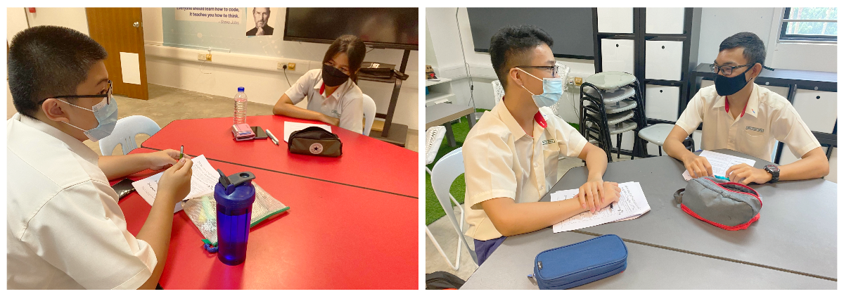 Four masked students in school uniforms sit at tables with notebooks and pencil cases in a classroom.