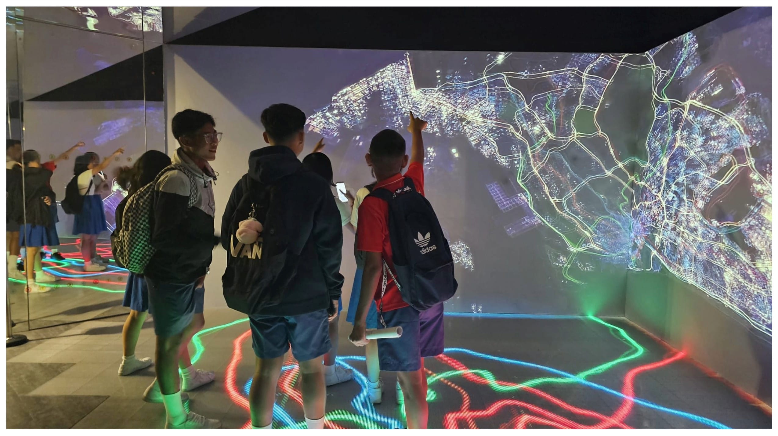 Children with backpacks point at a large wall projection of a city map, standing on a floor with colorful light lines.