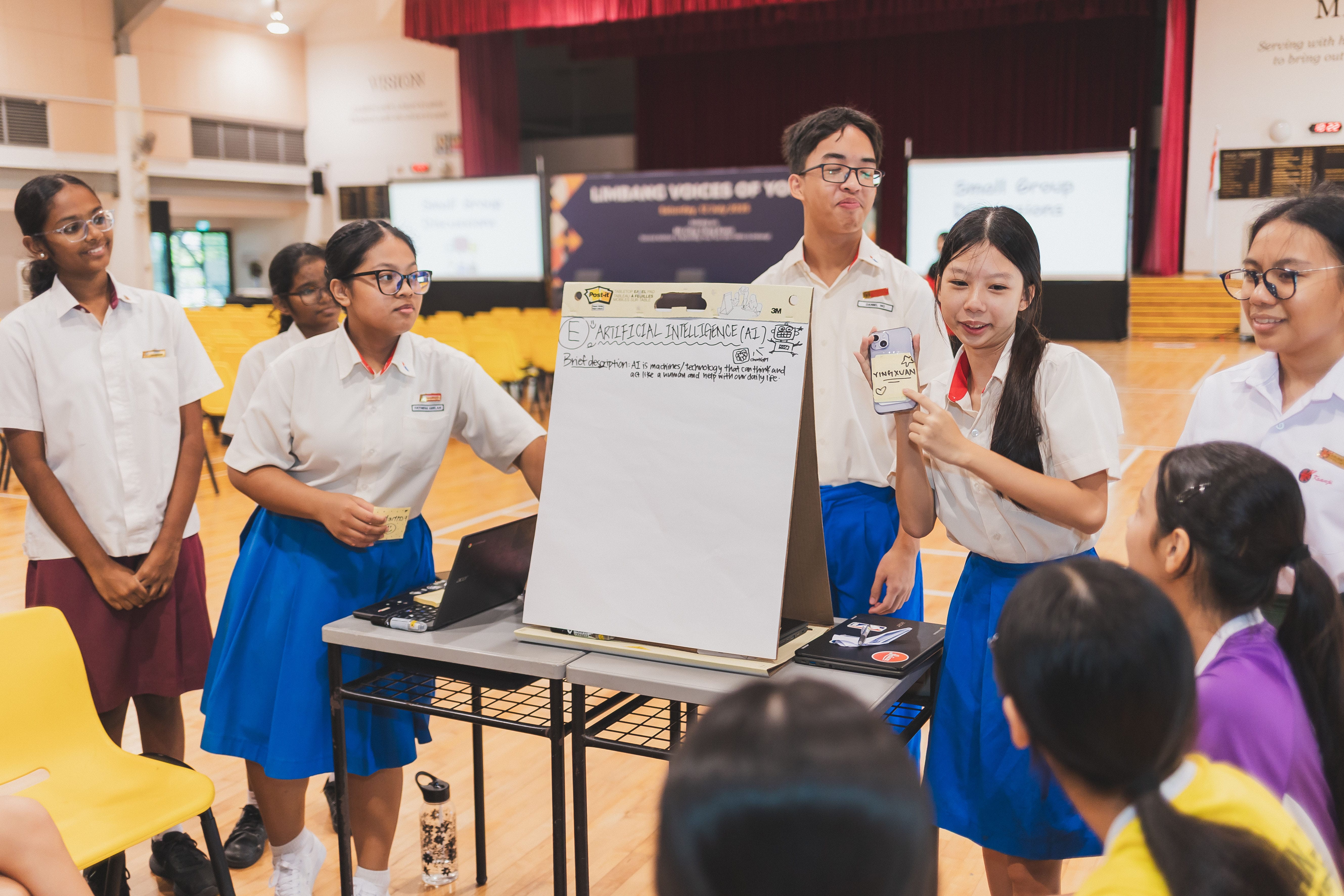 Students in school uniforms present a whiteboard about Artificial Intelligence in a school hall.