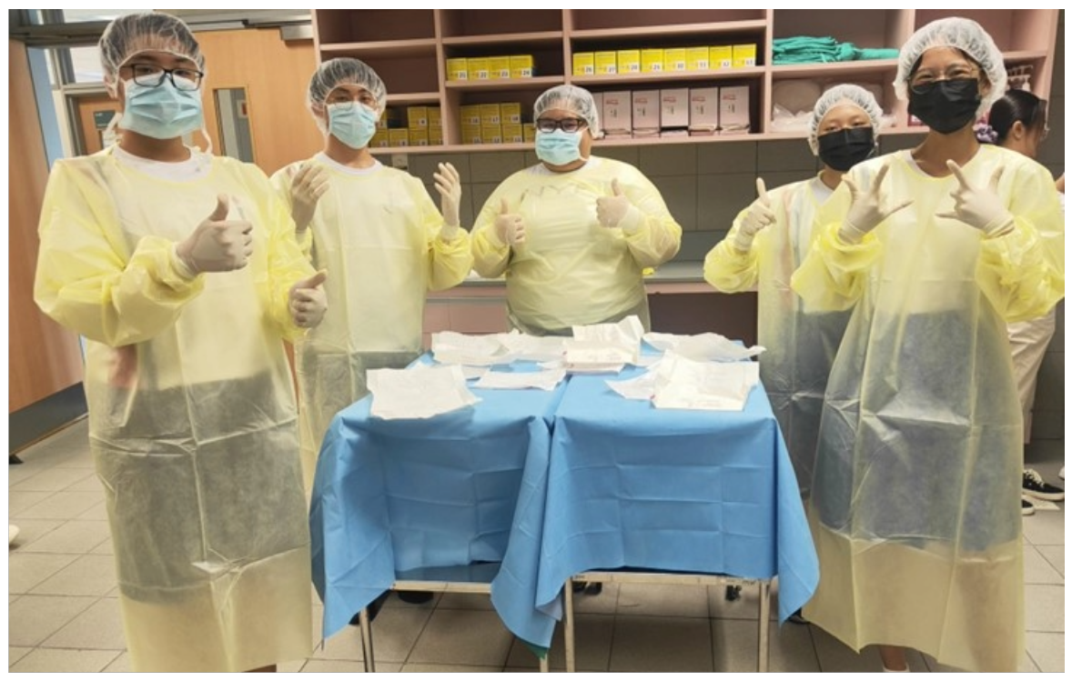 Five people in yellow gowns, gloves, hair nets, and face masks give thumbs up around a blue-draped table.
