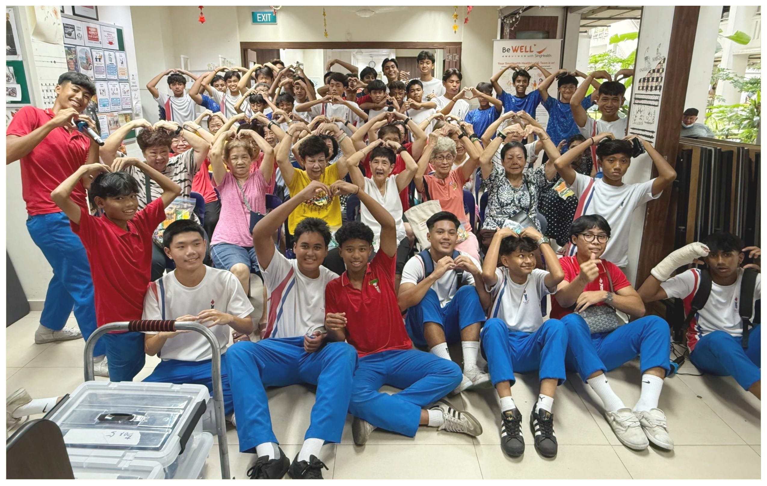 A large group of students and elderly people pose together indoors, making heart shapes with their hands overhead.