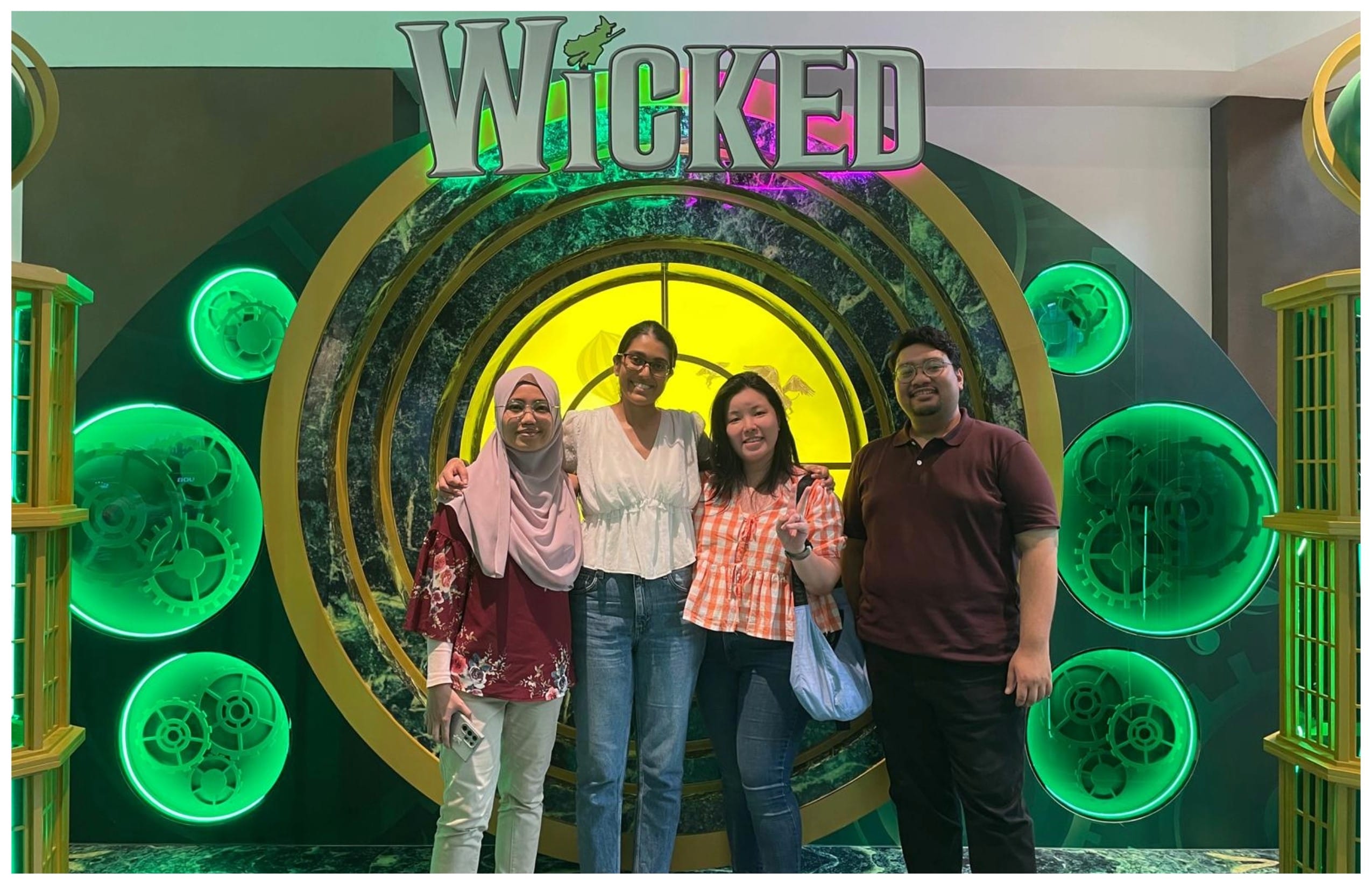 Four people pose in front of a green and gold Wicked musical display with glowing gear decorations.