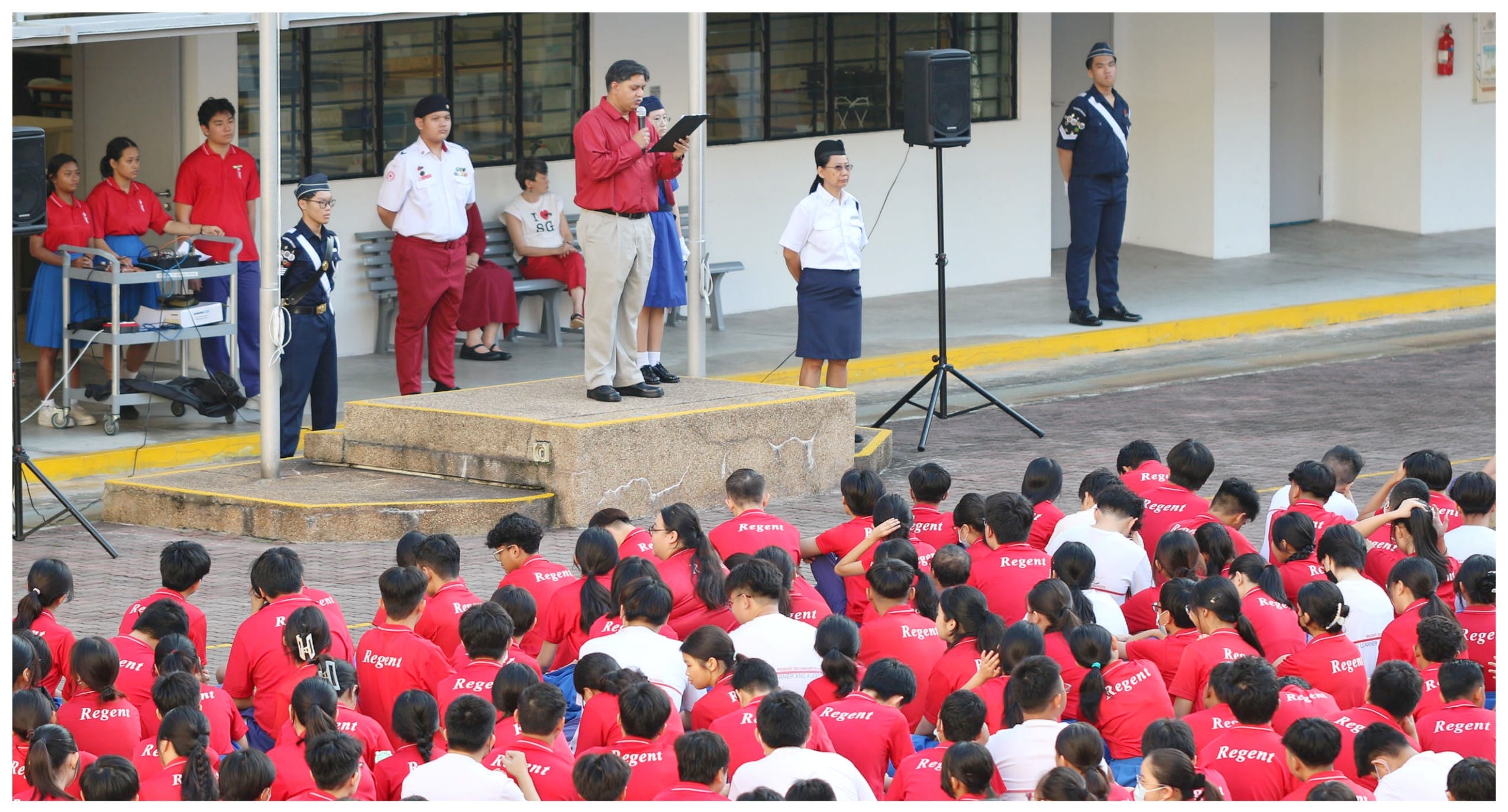 A man speaks at a school assembly while students in red Regent shirts sit on the ground facing a stage.